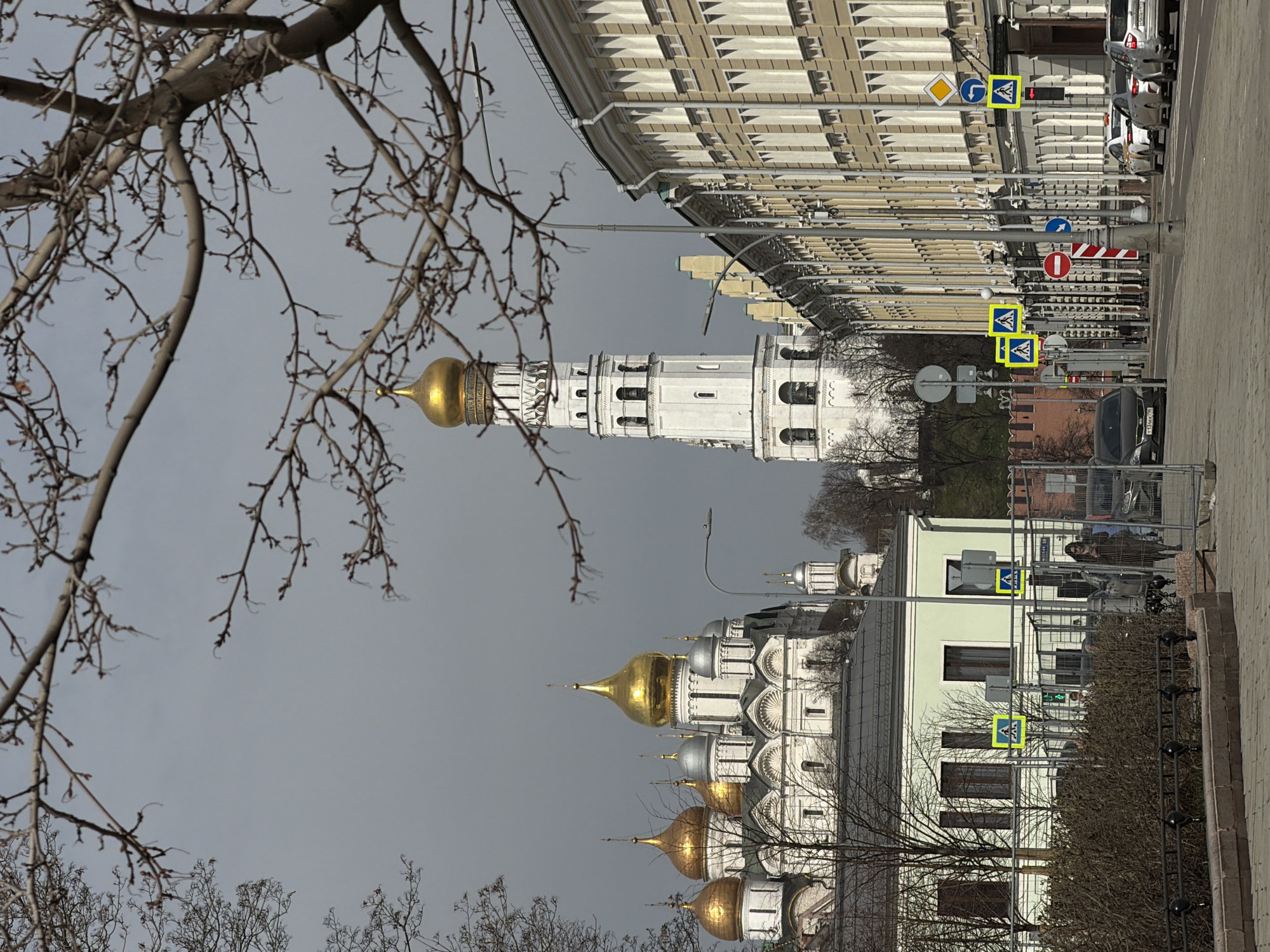 The image captures a Moscow streetscape featuring the Moscow Kremlin. In the foreground, bare tree branches frame the view of several prominent structures. Notably, a white bell tower and a church with golden domes are visible, typical features of the Kremlin's architectural ensemble. The buildings stand along a street equipped with pedestrian crossing signs. Further down the street, a long building stretches into the distance, with cars parked along the side. The sky is overcast.