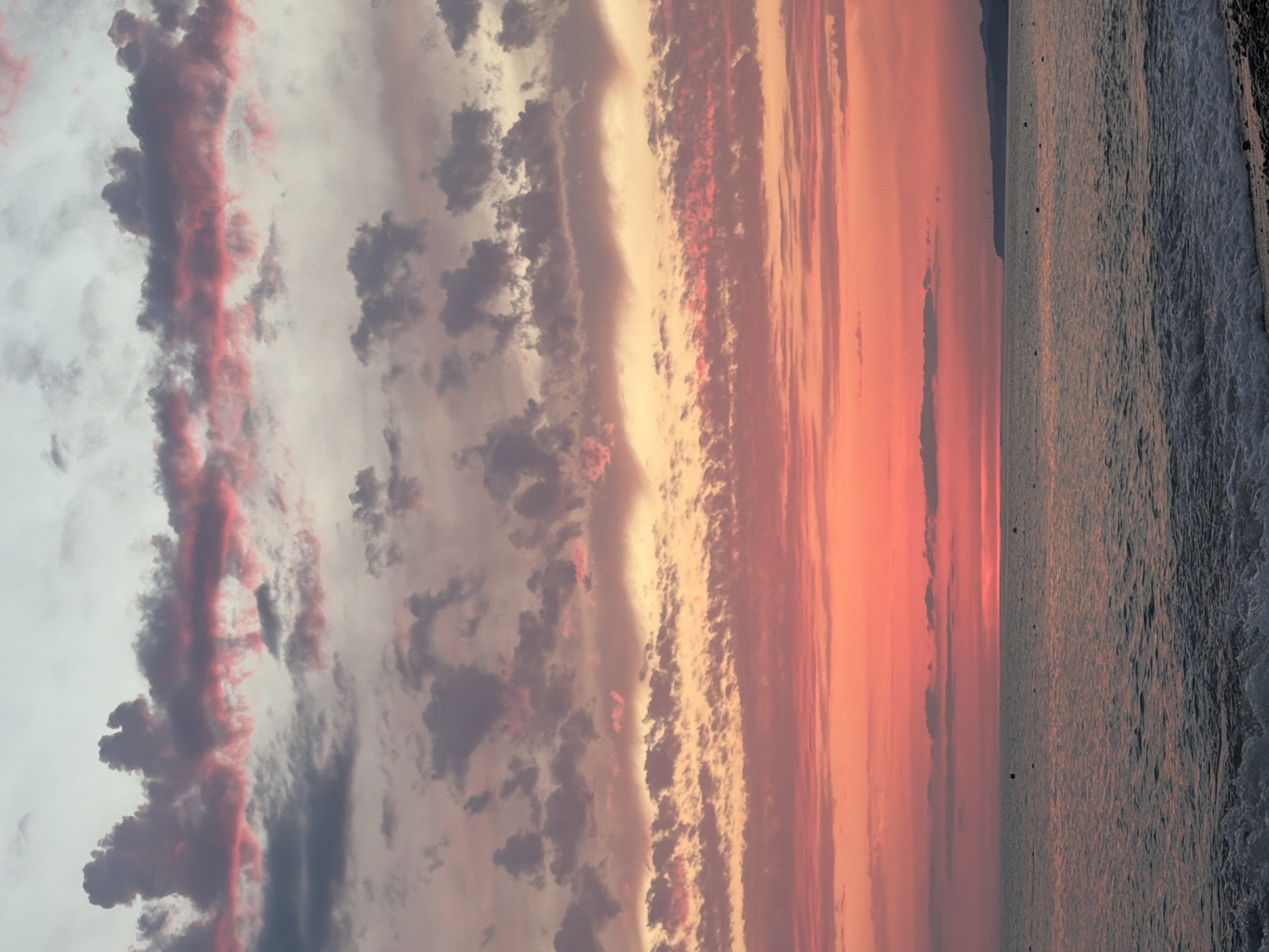 The image shows a picturesque sunset over the sea. The sky is a vibrant canvas of colors, ranging from deep reds and oranges at the horizon to soft pinks and purples higher up, transitioning into a pale blue at the very top. Wispy clouds are scattered throughout, catching the sunset light and adding texture to the sky. The sea is calm, with gentle waves lapping at the shore. The shoreline is composed of smooth pebbles. In the distance, a faint silhouette of land can be seen. The overall impression is one of a peaceful and serene evening by the sea.