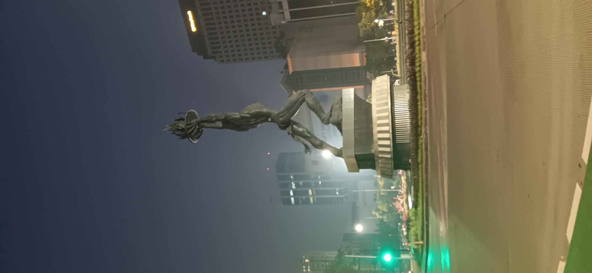The image depicts the Selamat Datang Monument (Welcome Monument) at night in South Jakarta, Indonesia. The monument, a bronze sculpture of a couple raising their arms, stands tall on a multi-tiered base in the center of a roundabout. The sky is dark and slightly hazy. Surrounding the monument are several tall buildings, including a prominent one with a "Maybank" logo illuminated at its top. Streetlights and traffic lights, including a green traffic light, are visible, casting a glow on the wet road surface, suggesting recent rain. Vegetation and some smaller structures are also present in the background. The overall scene captures a well-known landmark in an urban setting at night.