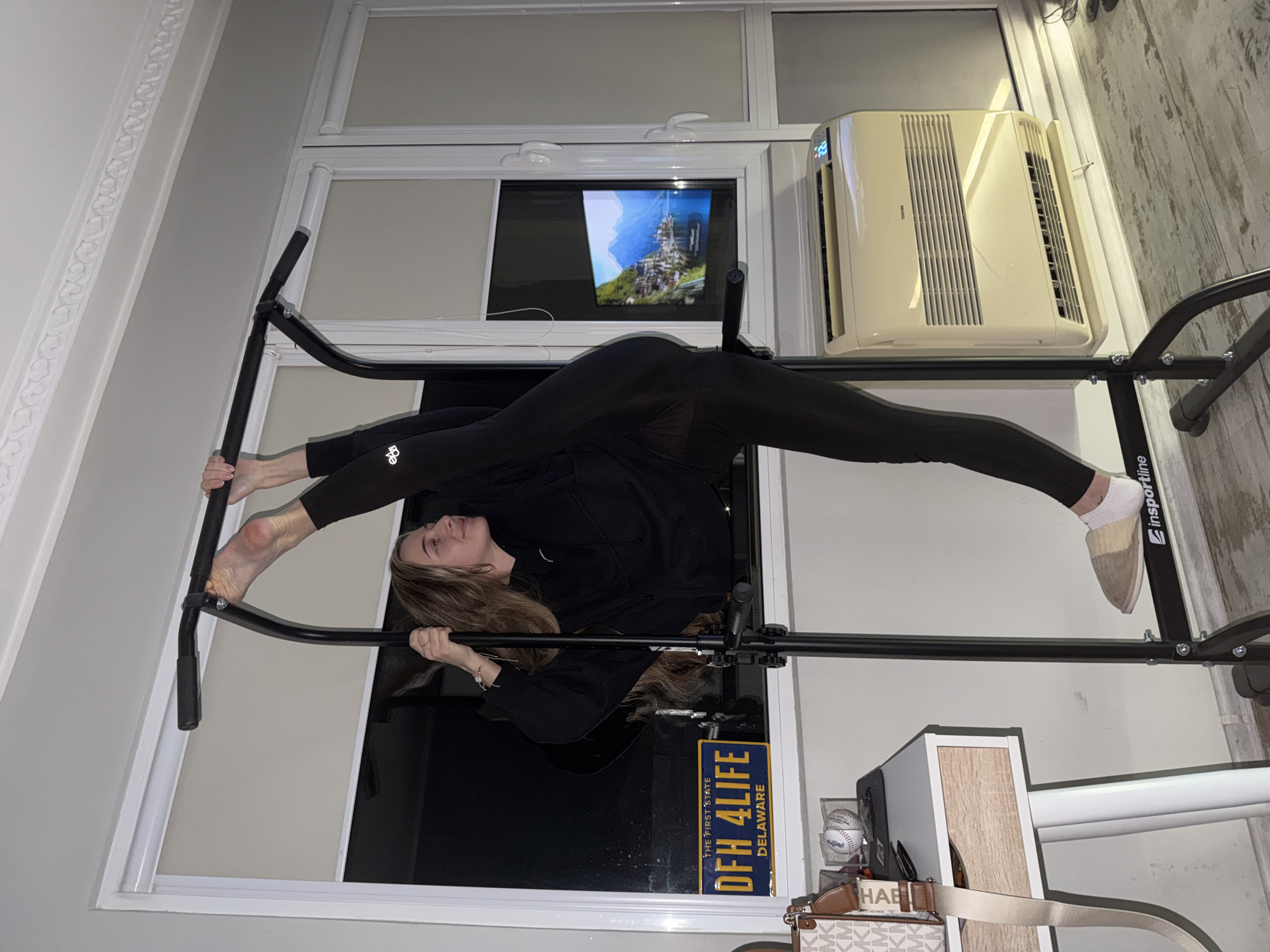 A woman in black leggings and a black sweatshirt is performing a split on a black pull-up bar inside an apartment. The pull-up bar is mounted in a window frame. Behind her, a flat-screen television displays an image of a castle. An air conditioner is mounted in the same window frame to the right of the television. Below the window, a desk with a purse, two baseballs, and a license plate that says "DFH 4 LIFE, Delaware" are visible. The walls are light gray. There is no evidence to suggest this image was taken in the city of Plovdiv, Bulgaria.