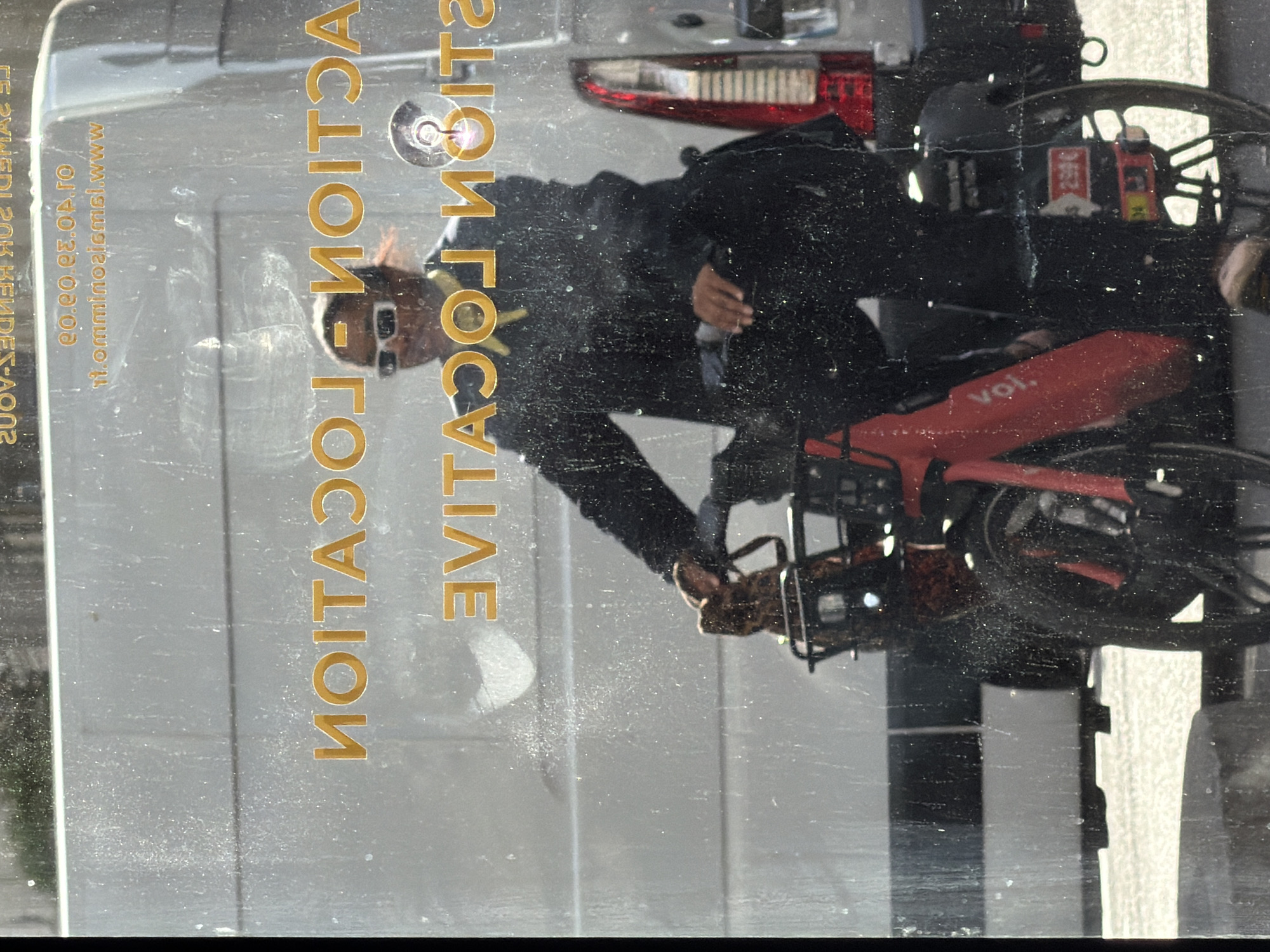 The image captures a person riding a red Voi-branded bicycle, likely in an urban setting during bright daylight. The photograph appears to have been taken through a reflective surface, possibly a window, resulting in a layered scene with prominent reflections and some dust or smudges.
The central subject is an individual riding the Voi bicycle. They are seen in profile, wearing dark clothing and sunglasses, with a light-colored or yellow band around their neck. The red bicycle features a front basket, which has a sticker displaying "250€" and possibly "34." The "voi." brand logo is visible on the bike's frame.
In the background, or reflected on the surface, is a white van with distinct red taillights, indicating the presence of other vehicles.
A prominent feature of the image is large, golden text reflected across the frame, which reads "ACTION LOCATION LIVE." Additional smaller, reversed text is visible on the reflective surface. This includes a website address, "it.omminioajcml.www," and a numerical sequence that appears to be a phone number: "00.00.00.00.00." Other partially visible and reversed text, "LE SVMEDE: SOK KEMDEE'S AOOS," is also present on the far left. The overall scene suggests an active street environment in Paris, France.