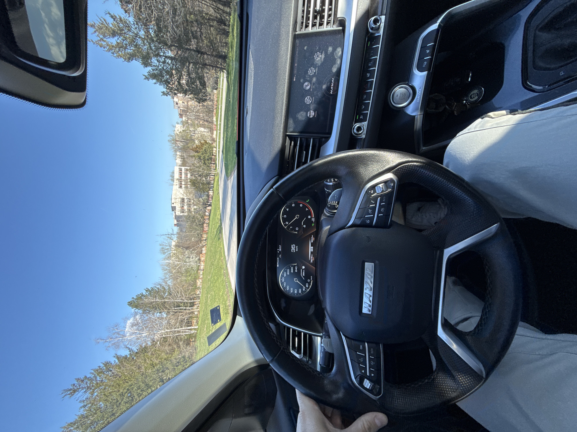 The image shows the interior of a car, specifically the dashboard, steering wheel, and a partial view of the driver's seat and the driver's hand. The steering wheel has the brand "HAVAL" on it. The speedometer reads 35 km/h. Through the windshield, a view of a clear blue sky, green lawn, trees, and buildings is visible. The scene potentially depicts a person driving a Haval vehicle in an area with open green spaces and buildings.
It is not possible to verify the location as Pleven, Bulgaria, based solely on the image. Additional evidence or context would be needed to confirm the location.