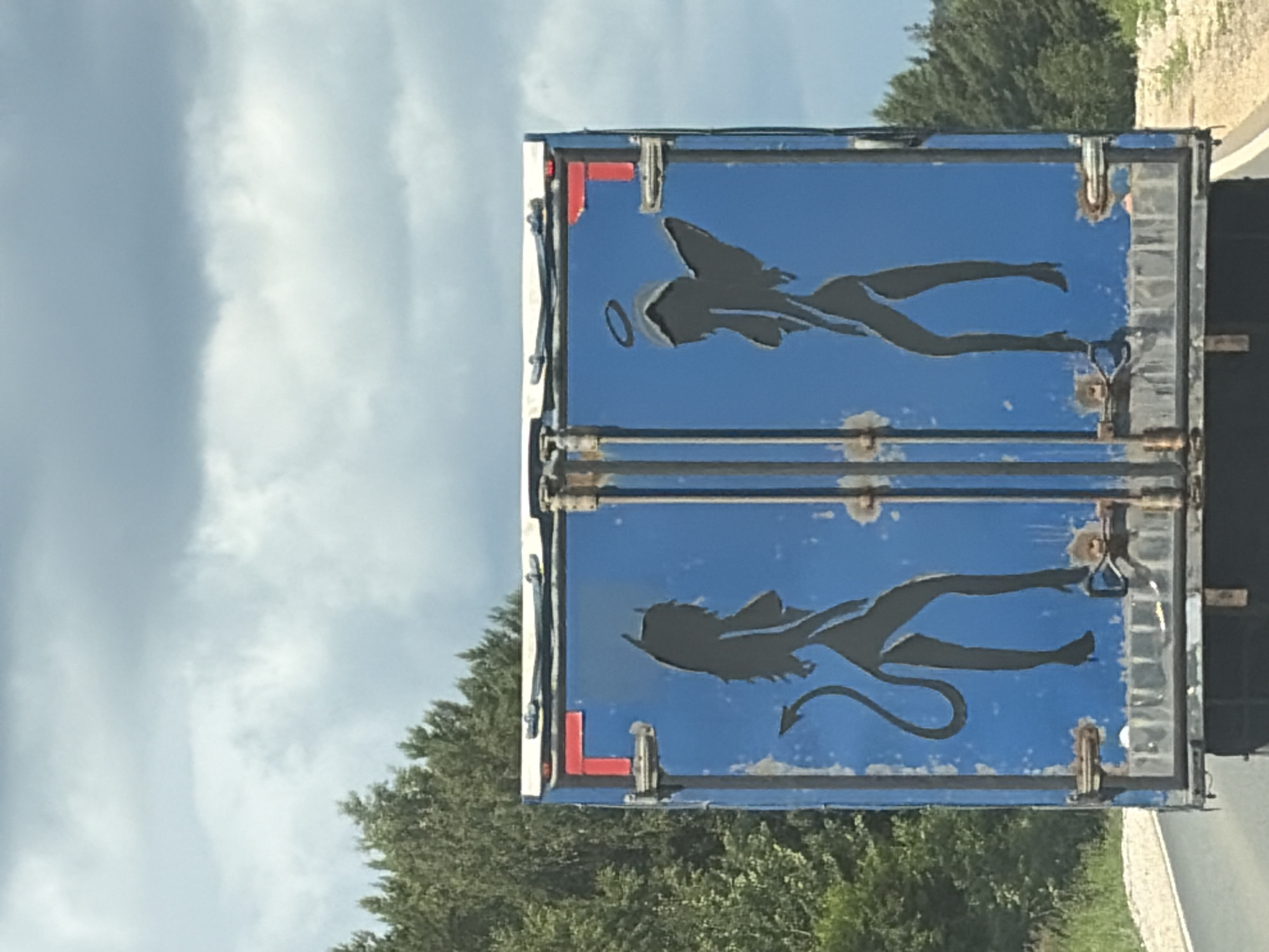 The image shows the back doors of a blue truck trailer with two black silhouette figures painted on it. On the left is a figure with a tail and horns, representing a devil. On the right is a figure with a halo and wings, representing an angel. The figures are stylized and appear to be in dancing poses. The trailer doors are weathered and show signs of rust and wear. The background consists of a cloudy sky and green trees, suggesting an outdoor setting, likely on a road given the perspective. The location is unknown.