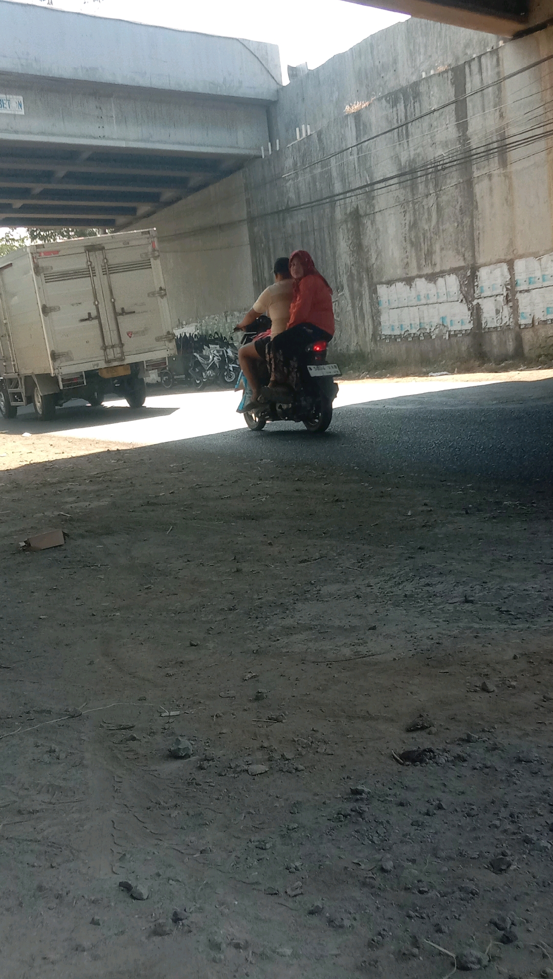 Two individuals are riding a motorcycle under an elevated concrete structure in an outdoor urban environment. The person in the rear is wearing a red hijab and an orange jacket. The person in front is wearing a tan shirt. The motorcycle is black and has a license plate that reads "B 3834 YAA". To the left of the motorcycle, a white box truck is parked. In the background, several other motorcycles are parked. The ground is covered in dirt and debris, and there is a large concrete wall to the right of the frame. The scene appears to be located on a street in Karang Anyar, Indonesia.