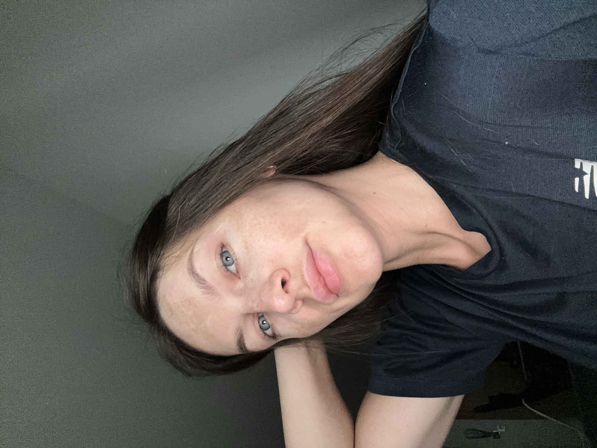 A young adult female with light skin, blue eyes, and brown hair is pictured in a close-up, angled selfie. Her head is tilted to the left, and she rests her left arm on the surface below, possibly a desk or table. She has noticeable freckles on her face. She is wearing a dark-colored t-shirt with white text/logo visible on the right side of her torso. The background is a plain, neutral-colored wall.
The image doesn't contain information or details that allow one to identify the location as Donji Pobori, Montenegro. The background and objects visible are non-specific and could be found in many indoor settings.