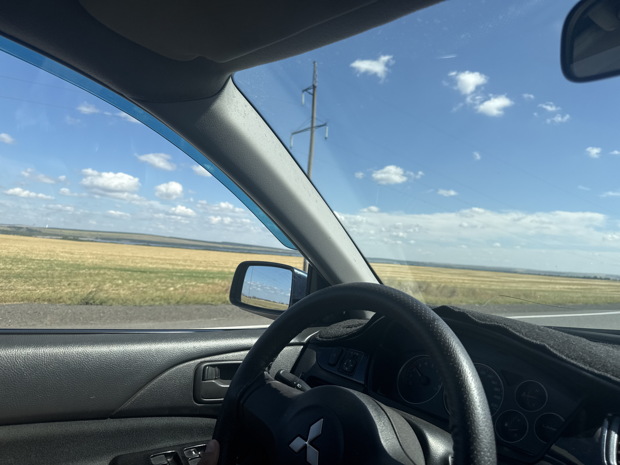 The image is a first-person view from inside a car, likely in the city of Sorochinsk, Russia, as indicated by the context. The foreground shows the interior of the car, with a black leather door panel, a steering wheel with the Mitsubishi logo, and a part of the dashboard with visible gauges and a gray dashboard mat. The driver's hand is partially visible on the steering wheel.
Through the windshield, a vast, flat landscape unfolds under a bright blue sky dotted with white clouds. The landscape appears to be agricultural, with fields of dry, yellowish-brown crops stretching to the horizon. A body of water, possibly a river or lake, is visible in the distance. A tall electricity pylon stands prominently against the sky, suggesting the presence of infrastructure in the rural area. A side mirror reflects the sky and a distant horizon. The overall impression is of a scenic drive through a countryside setting on a clear day.