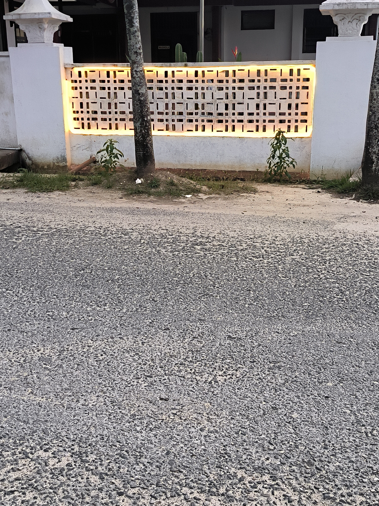 The image depicts a section of asphalt road in Langsa, Indonesia, bordering a low white wall. The wall is adorned with a decorative, latticed rectangular panel illuminated from behind with a warm yellow light. A slender tree trunk partially obscures the view of the panel's center. On the ground between the wall and the road, there is sparse vegetation, including small plants. The asphalt is a light gray color, characterized by a rough, aggregate texture. The background shows a building with dark windows and a partially visible sign stating "MUSHOLA". The lighting suggests a time of day close to dusk or dawn. There is no apparent human activity in the scene.