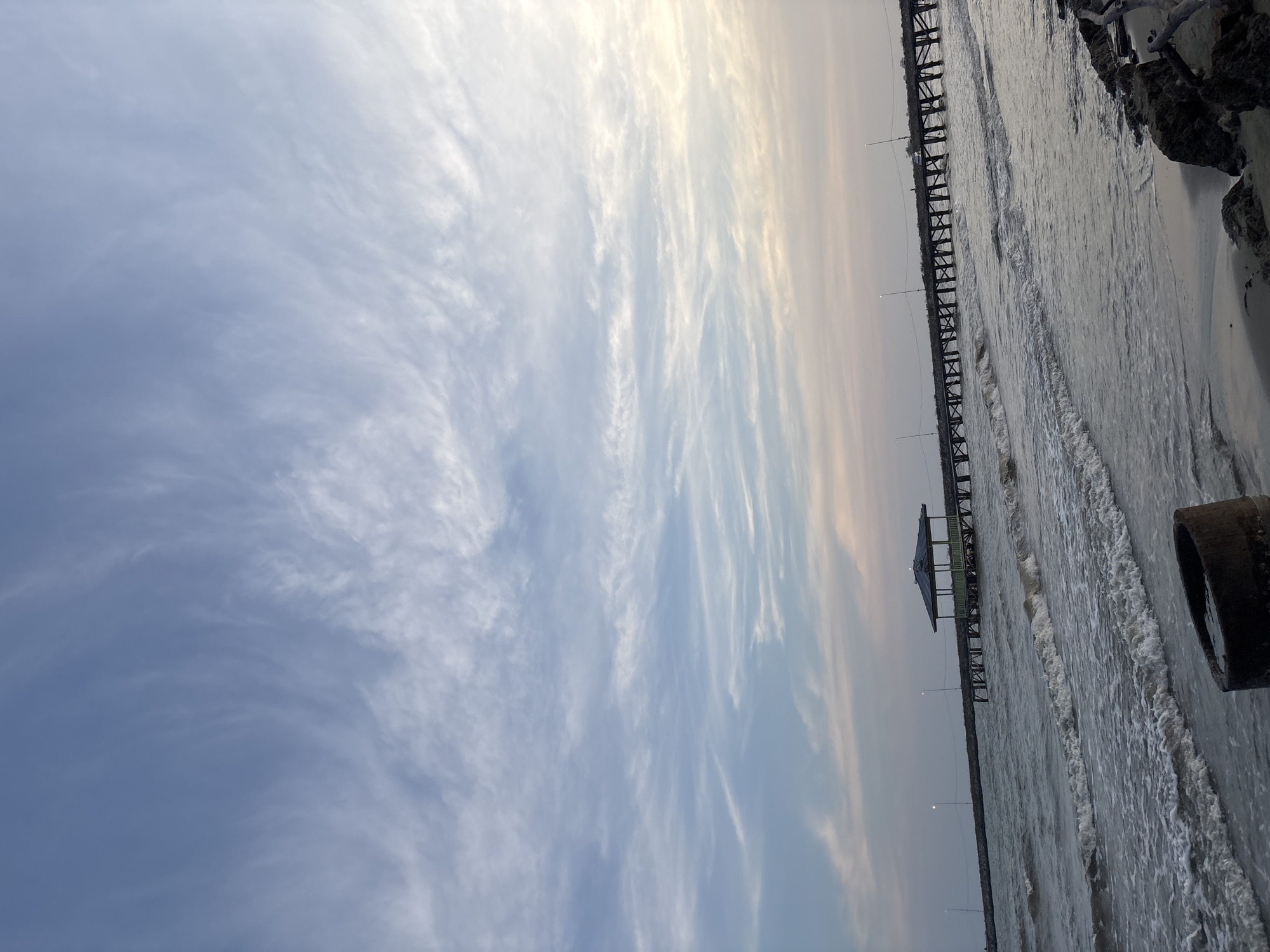 The image depicts a serene beach scene in Torosiaje Jaya, Indonesia, under a vast, cloud-streaked sky. A long, wooden pier stretches out into the ocean, supporting a small, open-air pavilion with a dark roof. The sea is moderately choppy, with waves gently rolling onto the sandy shore. In the foreground, a section of a concrete pipe lies on the beach, partially submerged in sand and water. Rocks and driftwood are scattered along the coastline. The sky displays a soft gradient of blue and pale pink, suggesting either early morning or late evening light. There are no people visible in the image.