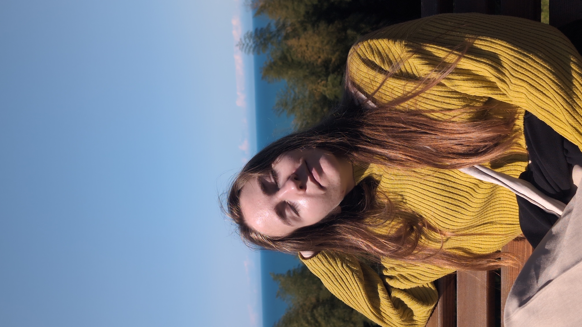 A woman is lying on a wooden bench outdoors. She has long brown hair and is wearing a mustard-yellow ribbed sweater. Her eyes are closed, and she appears to be resting. The background includes a view of the sky, a body of water that could be the Black Sea, and some trees. The location is likely in Batumi, Georgia, given the prompt's specification.