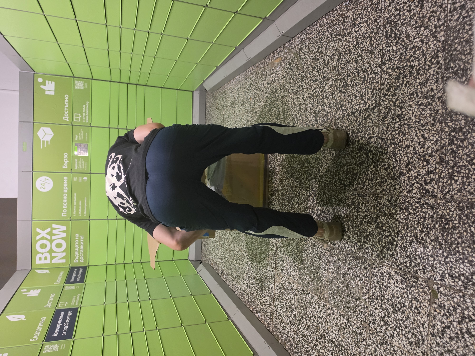 A person, viewed from behind, wearing a black t-shirt and dark blue track pants with a white stripe, is bent over and appears to be interacting with a large cardboard box at a BOX NOW automated parcel locker station. The locker station, painted bright green, is visible in the background with various text and icons on its surface. Text in Bulgarian on the lockers reads "BOX NOW", "Бъдещето на доставките!" (The future of deliveries!), "24/7", "По всяко време" (At any time), "Бързо" (Fast), and "Достъпно" (Accessible). The floor is a speckled terrazzo. The scene suggests a commercial or public location, likely within Sofia, Bulgaria, given the Bulgarian language text.