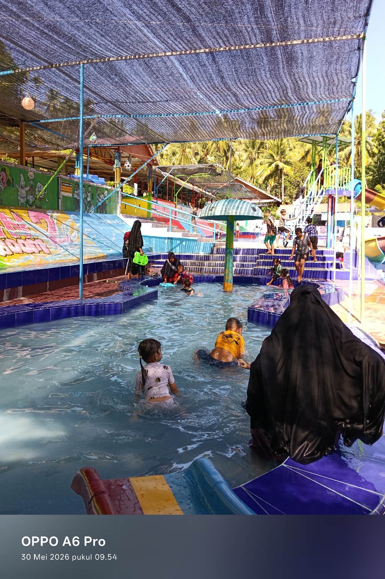 The image depicts a group of people enjoying a pool at what appears to be a water park. The location is likely in Palohseulimeng, Indonesia. Several children are in the pool, some are swimming, and others are standing or wading in the water. There are adults present, some of whom are wearing hijabs. In the background, there are colorful slides, stairs, and other water park features. A mesh-like structure provides shade over the pool area. The photograph was taken with an OPPO A6 Pro on May 30, 2026, at 09:54 AM.