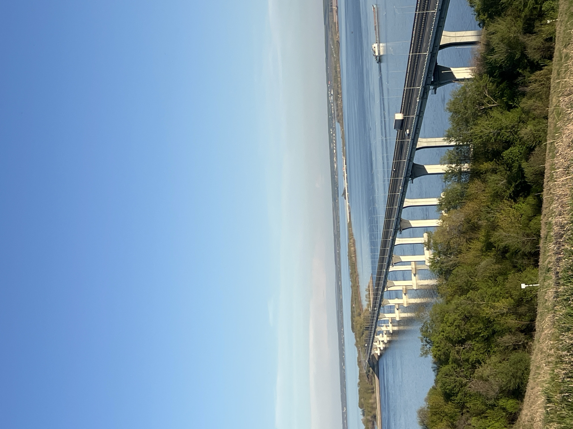 The image depicts the Sorochyi Hory Bridge spanning the Kama River near Сорочьи Горы, Russia. The bridge is a multi-span concrete structure with a roadway supported by piers. Water surrounds the bridge, indicating the river's presence. On either side of the bridge approach, there are strips of land and vegetation. A small barge is seen under the bridge. The sky above is clear blue with some light clouds. The photograph is taken at an unusual angle, resulting in a rotated perspective. A vehicle is visible on the bridge roadway.