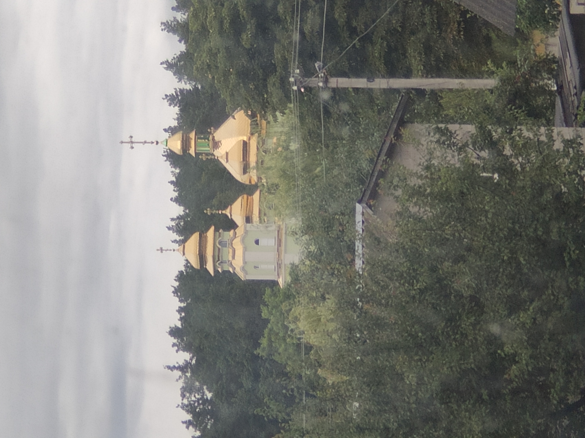 The image shows a view of a church with golden domes behind a dense canopy of green trees. The sky is overcast and gray. In the foreground, to the right of the church, a concrete utility pole with wires is visible. To the lower left of the image, a portion of a building with a slanted roof is also visible, along with more trees. The overall impression is of a church situated in a somewhat rural or suburban area with significant vegetation. The church has multiple domes, with at least two prominent golden domes and a smaller one visible to the left. The church structure appears to be light-colored, possibly white or pale green. The image is taken from a distance, and the details are somewhat obscured by the foliage and the distance. There are no people visible in the image.