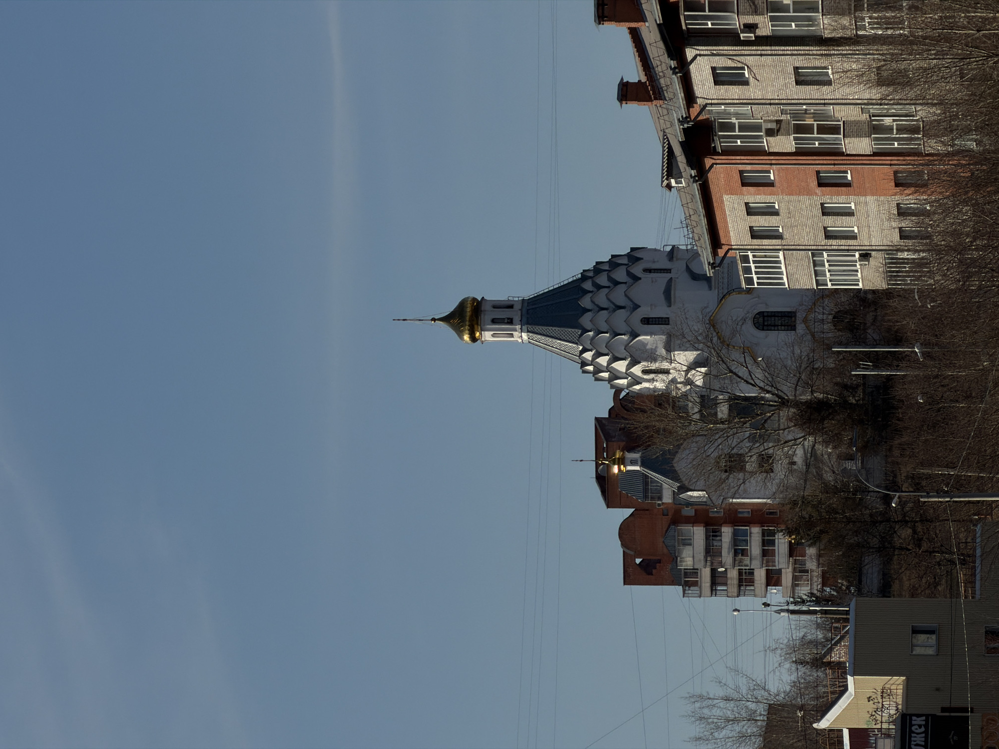The image shows a building located in Krasnoyarsk, Russia. The building has a white facade with a blue roof and a golden spire on top. There are several other buildings nearby, some with brick facades and some with grey facades. There are trees in the foreground, and a clear blue sky in the background. There are overhead wires running through the sky. The image has been rotated 90 degrees counter-clockwise.