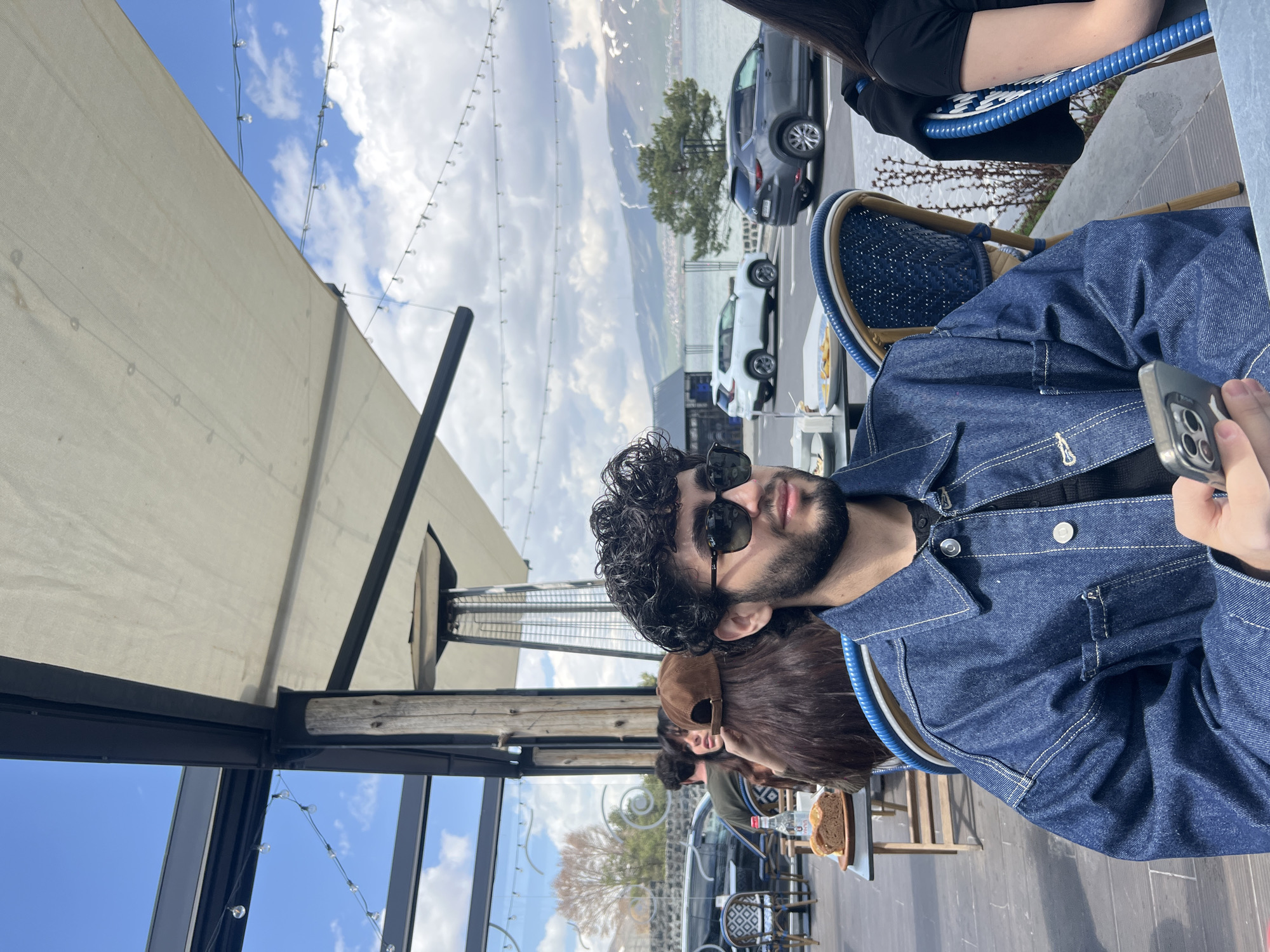 The image captures a scene at an outdoor cafe in an unknown city. A man with dark curly hair and a beard is wearing sunglasses and a denim jacket. He is sitting at a table and holding a smartphone. Several other people are also seated at nearby tables. Through the cafe's glass enclosure, cars and trees are visible in the distance, along with a body of water and hills under a partly cloudy sky. Overhead, a beige awning stretches across the top of the outdoor seating area, with string lights hanging beneath it.