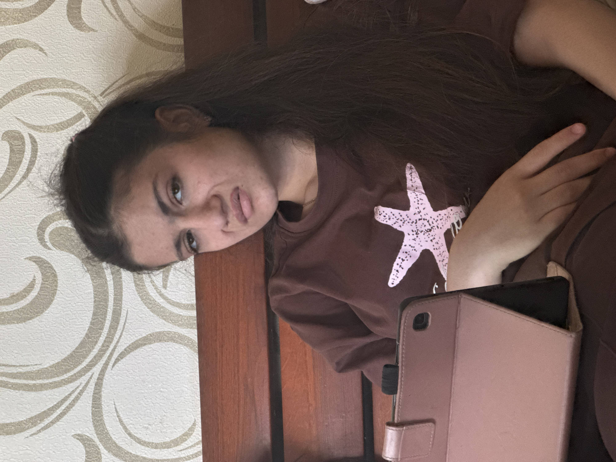 A single female individual is depicted indoors, likely in a bedroom, in the city of Unknown, Unknown. She possesses long, dark brown hair, partially styled up with some strands loose, and is wearing a brown long-sleeved shirt featuring a large, light pink starfish graphic on the chest. Her facial expression is a grimace, with pursed, downturned lips and slightly furrowed brows, indicating disgust or displeasure as she looks directly towards the camera. She has light skin and a faint gold chain visible around her neck, with an earring in her visible ear. She appears to be reclining against a dark brown wooden headboard, which has horizontal slats. In front of her, a tablet in a pink or rose gold protective case is propped upright, supported by its integrated stand. A black elastic band is wrapped around the top of the tablet case. The background consists of cream-colored wallpaper with a repeating pattern of large, swirling, abstract designs in a light brown or beige hue. Her hands are positioned near the tablet and her side.