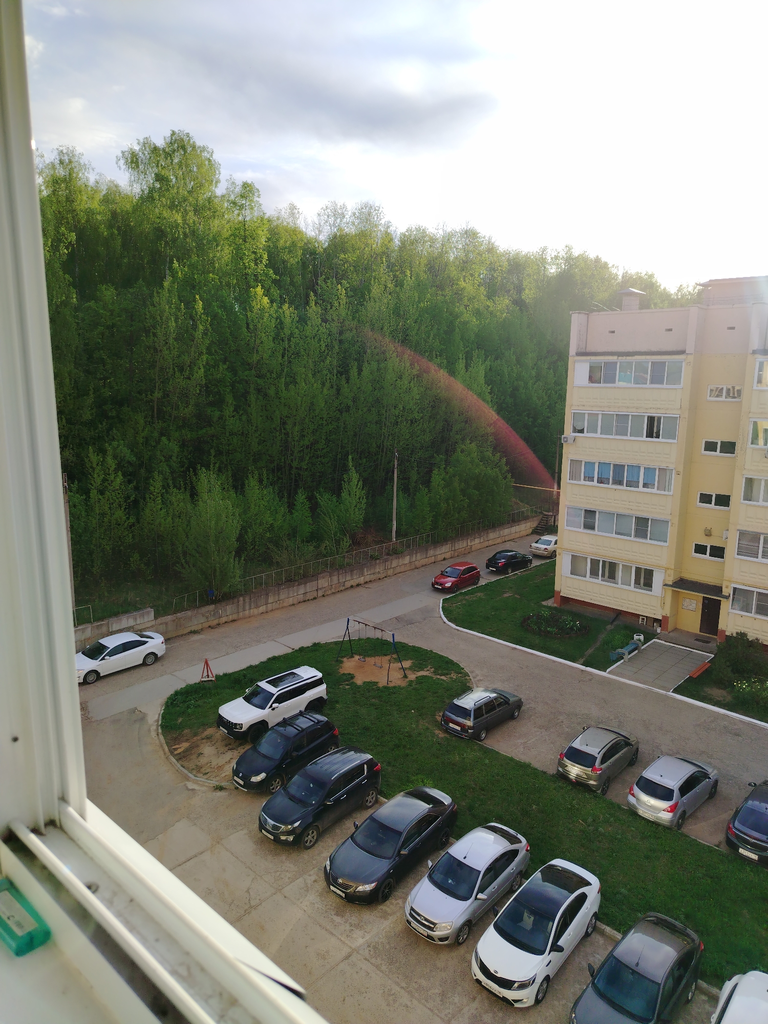 The image presents an elevated view, likely from a window, overlooking a residential area in Noviy, Russia. The scene includes a five-story yellow apartment building adjacent to a dense green forest. A paved road and parking area are visible, populated by approximately 14 parked cars of various makes and models. There is a small children's playground with swings. A red arc resembling a sprinkler or water source appears between the trees and the building. The sky is overcast with sunlight breaking through, suggesting it is daytime. The environment is urban with a strong element of nature.