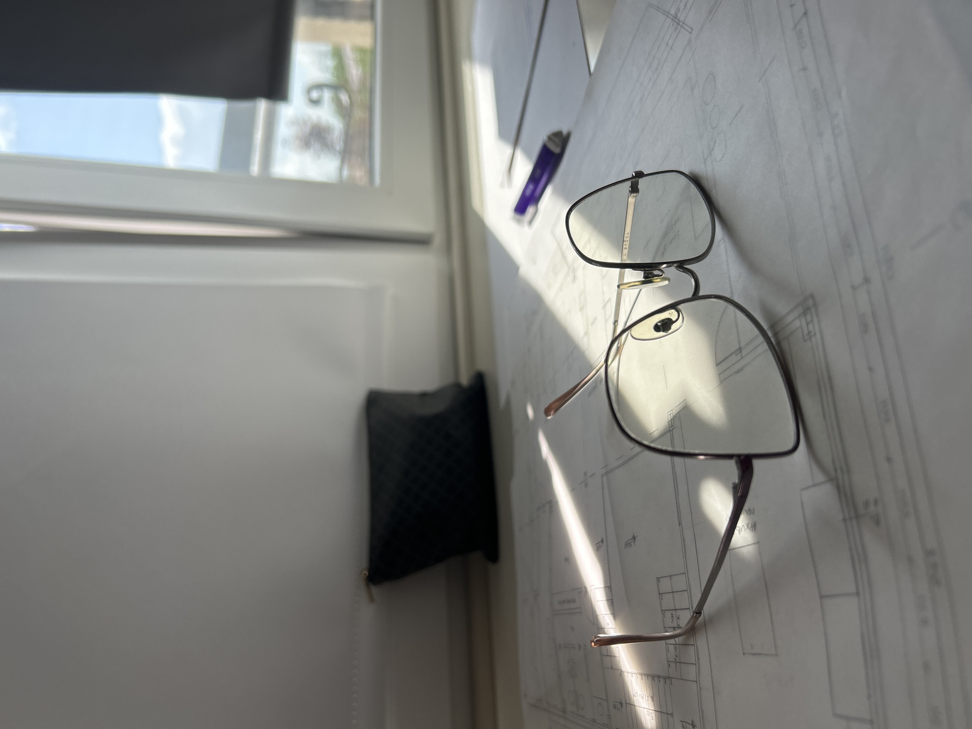 The image shows a pair of glasses resting on a large sheet of paper with architectural drawings. A purple pen lies next to the glasses, and a black, quilted pouch hangs against a white wall. Natural light illuminates the objects from a window, which reveals a view of the sky and trees. The setting appears to be an indoor space, possibly an office or studio. There is no indication that the location is in Plovdiv, Bulgaria; information about the city is not discernible from the image.