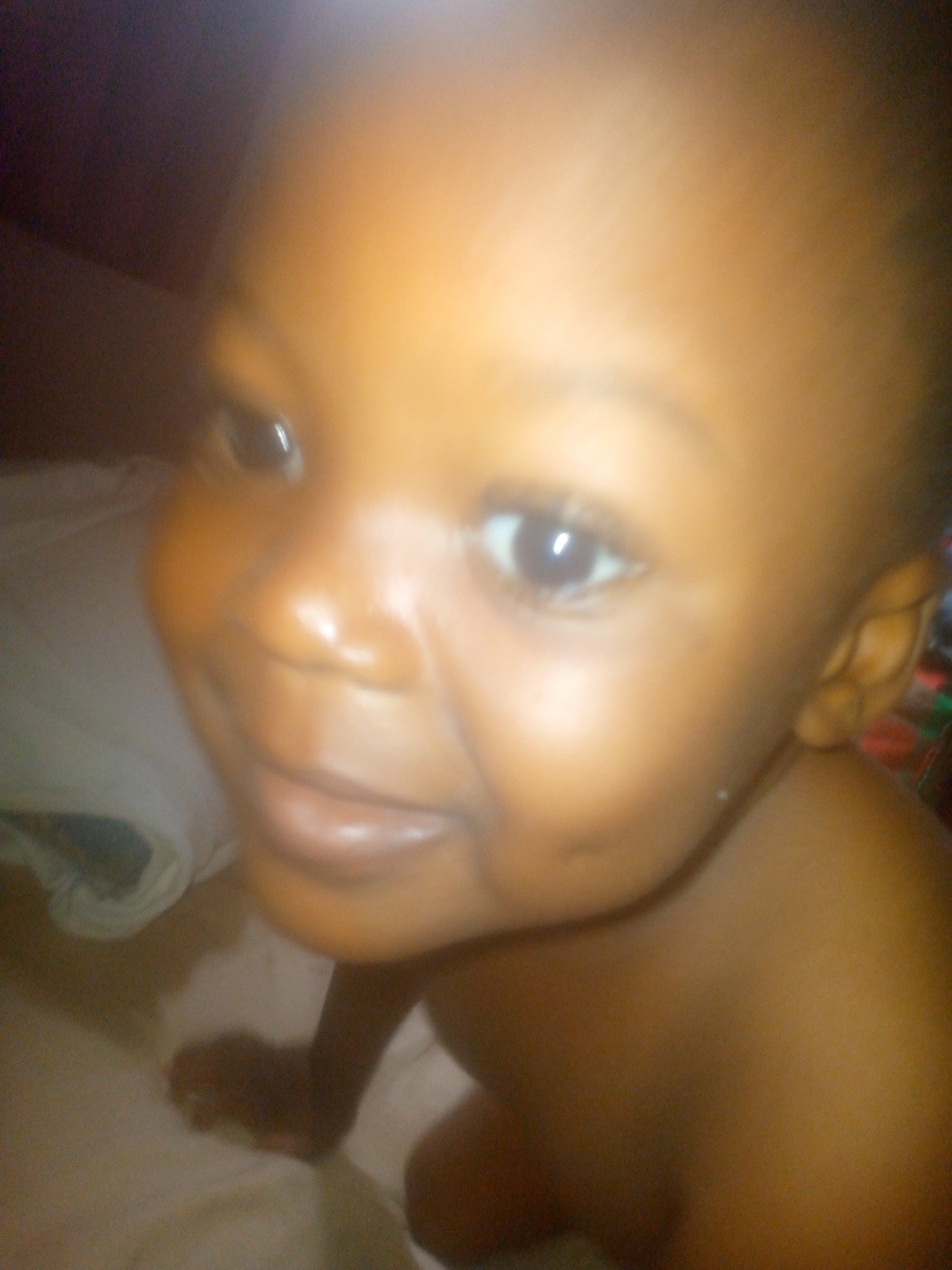 A close-up shot of a baby with dark skin. The baby is lying down, and their face is the main focus of the image. The background is blurry. The location is unknown.
