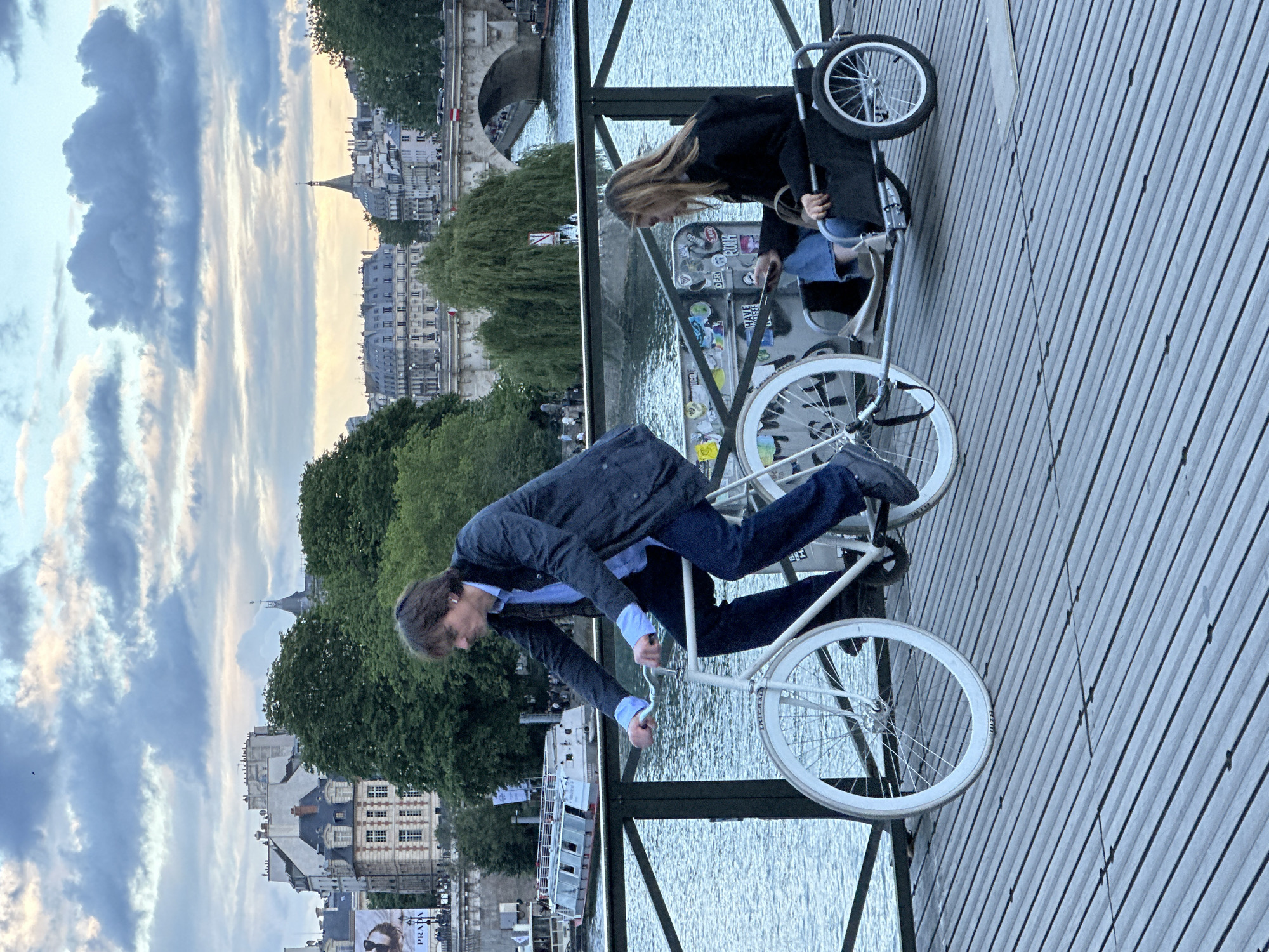 A man in a blue jacket rides a bicycle on the Pont des Arts bridge in Paris, France. A woman in a black jacket sits in a wheelchair nearby. The Seine river flows beneath the bridge, and buildings are visible in the background under a cloudy sky.