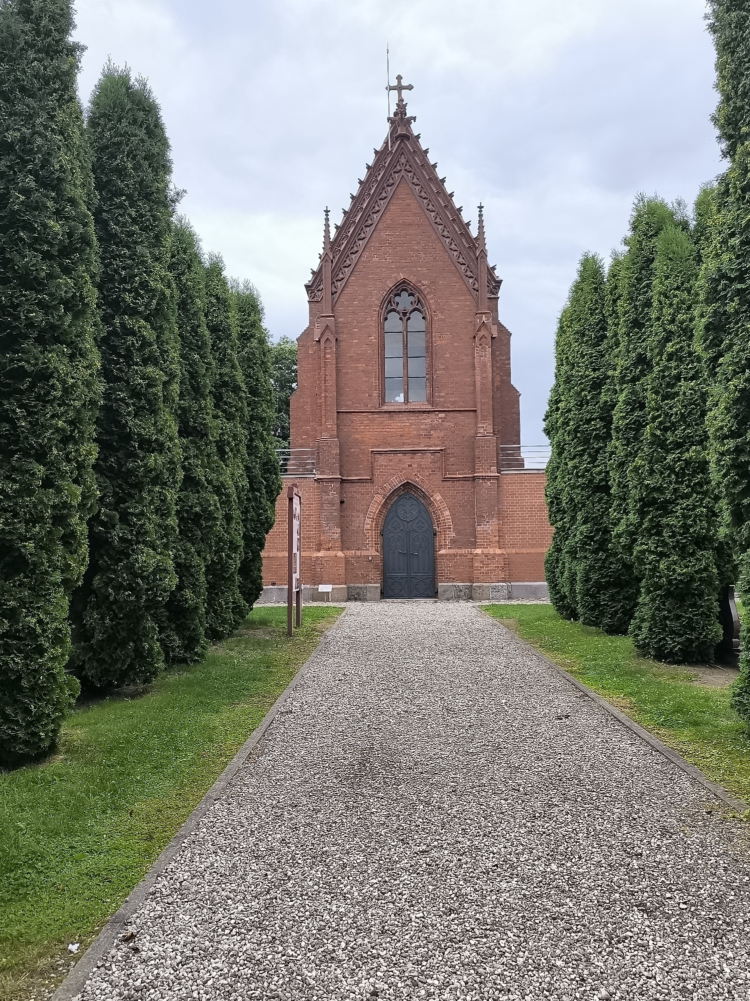 A gravel path leads towards a small, ornate red brick building, possibly a chapel or mausoleum, in Kretinga, Lithuania. The building features gothic architectural elements, including a steep, pointed roof with decorative pinnacles and a cross at its apex. A large, dark arched door is centrally positioned on the facade, above which is a tall, arched window with mullions. The building is flanked by rows of tall, conical evergreen trees, creating a symmetrical and formal entrance. A signpost stands to the left of the path. The sky is overcast.