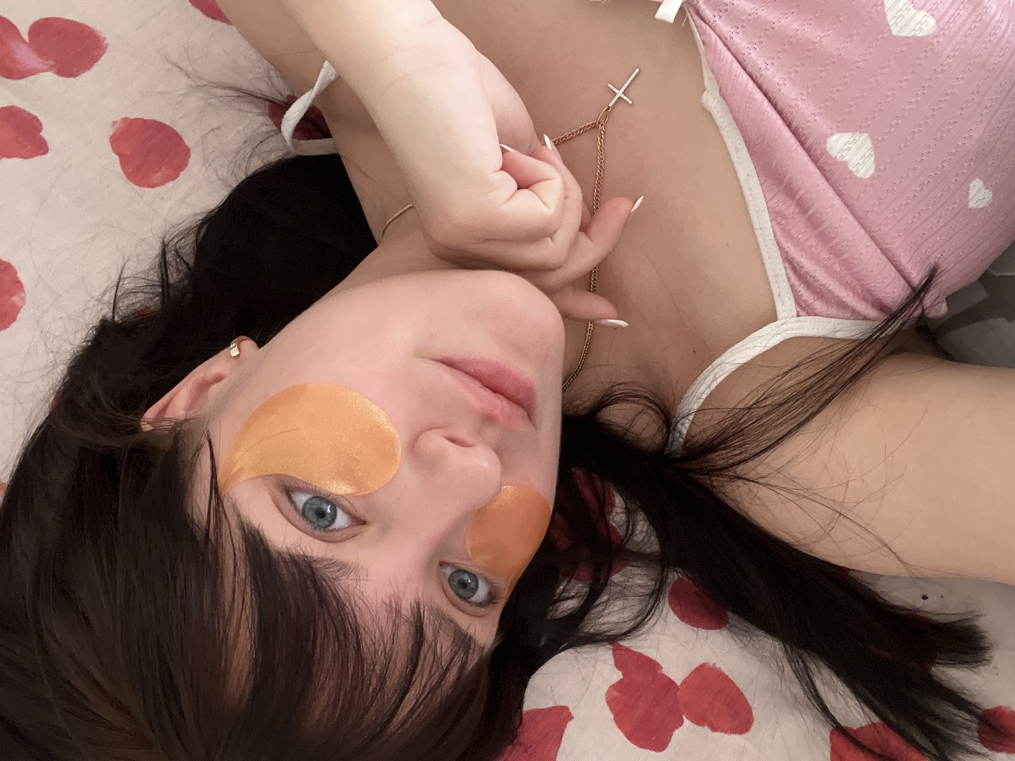 An overhead shot captures a young woman lying on her back on a bed, looking directly at the viewer. She has dark hair, which is spread around her head on the pillow. Her eyes are blue, and she wears golden-orange eye patches, two under her eyes and one on her forehead, indicating a self-care or beauty routine.
She is dressed in a light pink top with white trim and white heart patterns, suggesting sleepwear or comfortable loungewear. A delicate, gold-colored necklace with a cross pendant adorns her neck, with one of her hands gently resting near it, showcasing manicured fingernails with white tips. The bedding beneath her is white with scattered red, petal-like or heart-like patterns.
The scene conveys a moment of relaxation or contemplation in an indoor setting, likely a bedroom. The light appears soft, suggesting an intimate and calm atmosphere. No visible text is present in the image. The location context provided is Марковский, Russia.