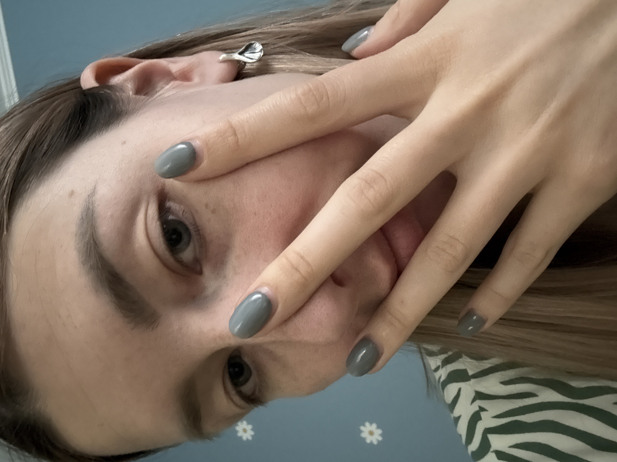 A close-up, tilted portrait of a woman is presented. The woman is light-skinned and has her hand partially covering her face. The nails on her hand are painted with a gray color. She has brown hair and gray eyes. An earring is visible on her left ear. The background features a blue surface with white daisy patterns. A zebra print clothing piece is visible near the bottom right corner. This image does not contain any information regarding the location in the city of Moscow, Russia.