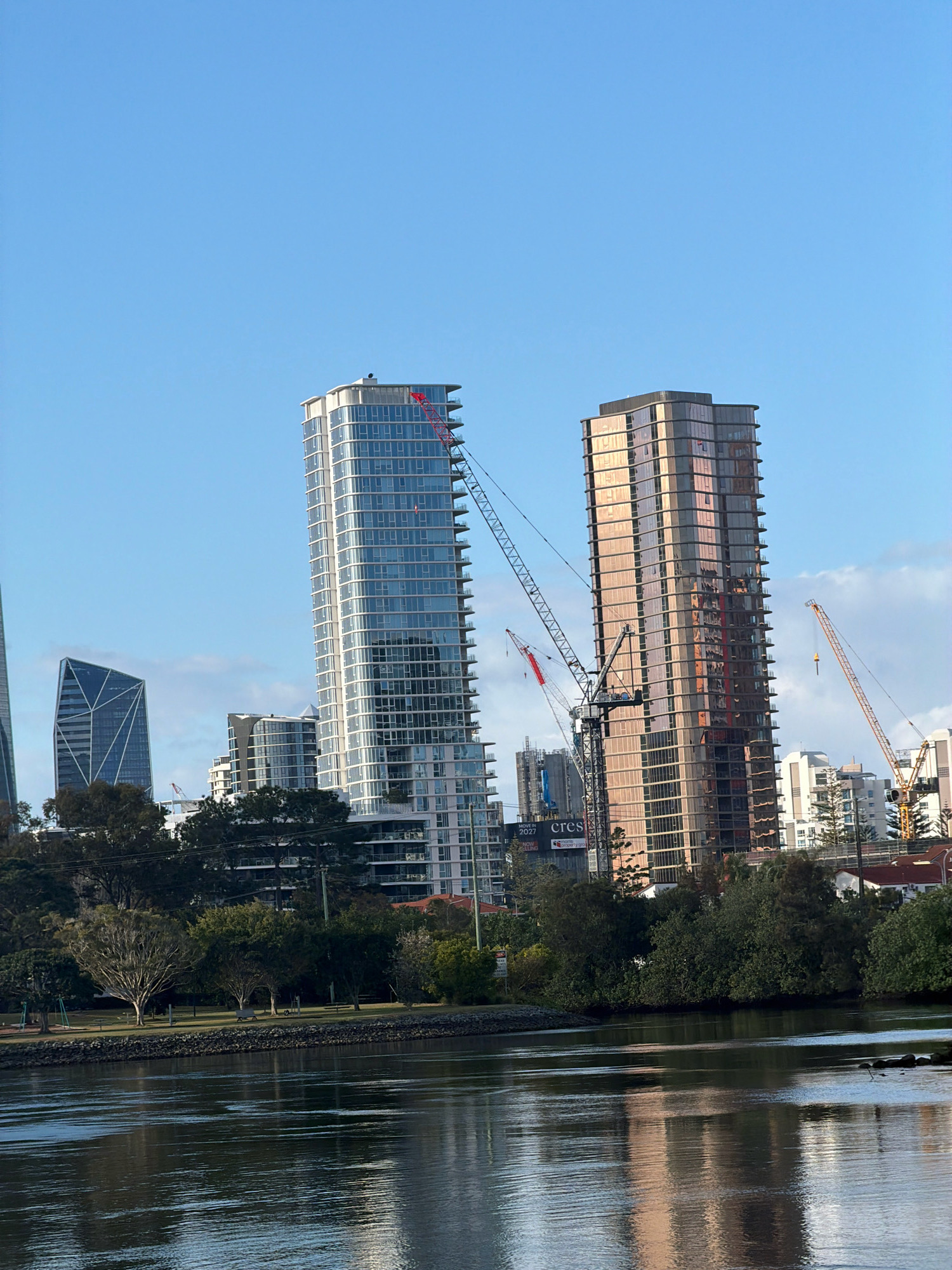 The image displays a waterfront view of Broadbeach Waters, Australia. In the foreground, a body of water reflects the surrounding environment, including the sky and buildings. Beyond the water, a park area with trees and grassy patches is visible. Several modern high-rise buildings dominate the background, some of which appear to be under construction, indicated by the presence of cranes. The sky is clear and blue, suggesting a daytime scene.