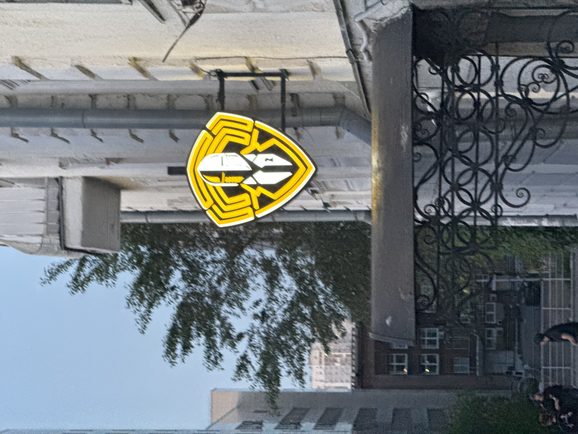 The image shows a close-up of a yellow and black shield-shaped logo with a stylized figure wearing a helmet and suit. The logo is mounted on the side of a building with light-colored textured walls and some vertical pipes. In the background, there are trees and other buildings, suggesting an urban environment. The lighting indicates it is either early morning or late evening. A dark metal awning with ornate scrollwork is visible in the foreground. On the street level, several blurry figures are visible, some standing and one appearing to be walking. The overall scene appears to be in a city, likely Yekaterinburg, Russia, based on the context.