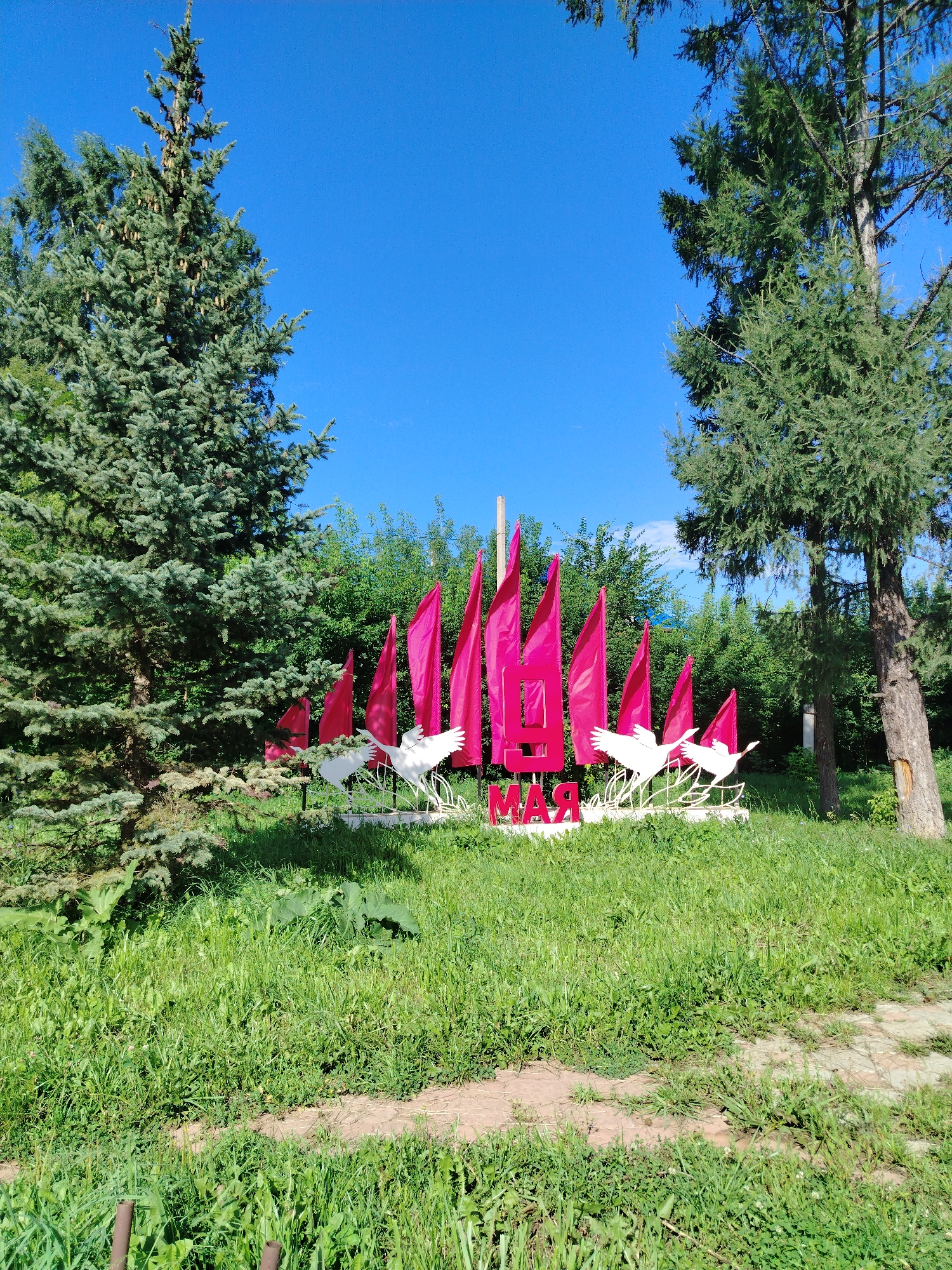 The image depicts a monument with red flags and white figures of birds in the city of Kuyeda, Russia. The monument is located in a grassy area with trees and blue sky in the background. The text on the monument appears to be in Cyrillic script. The overall scene suggests a festive or commemorative occasion.