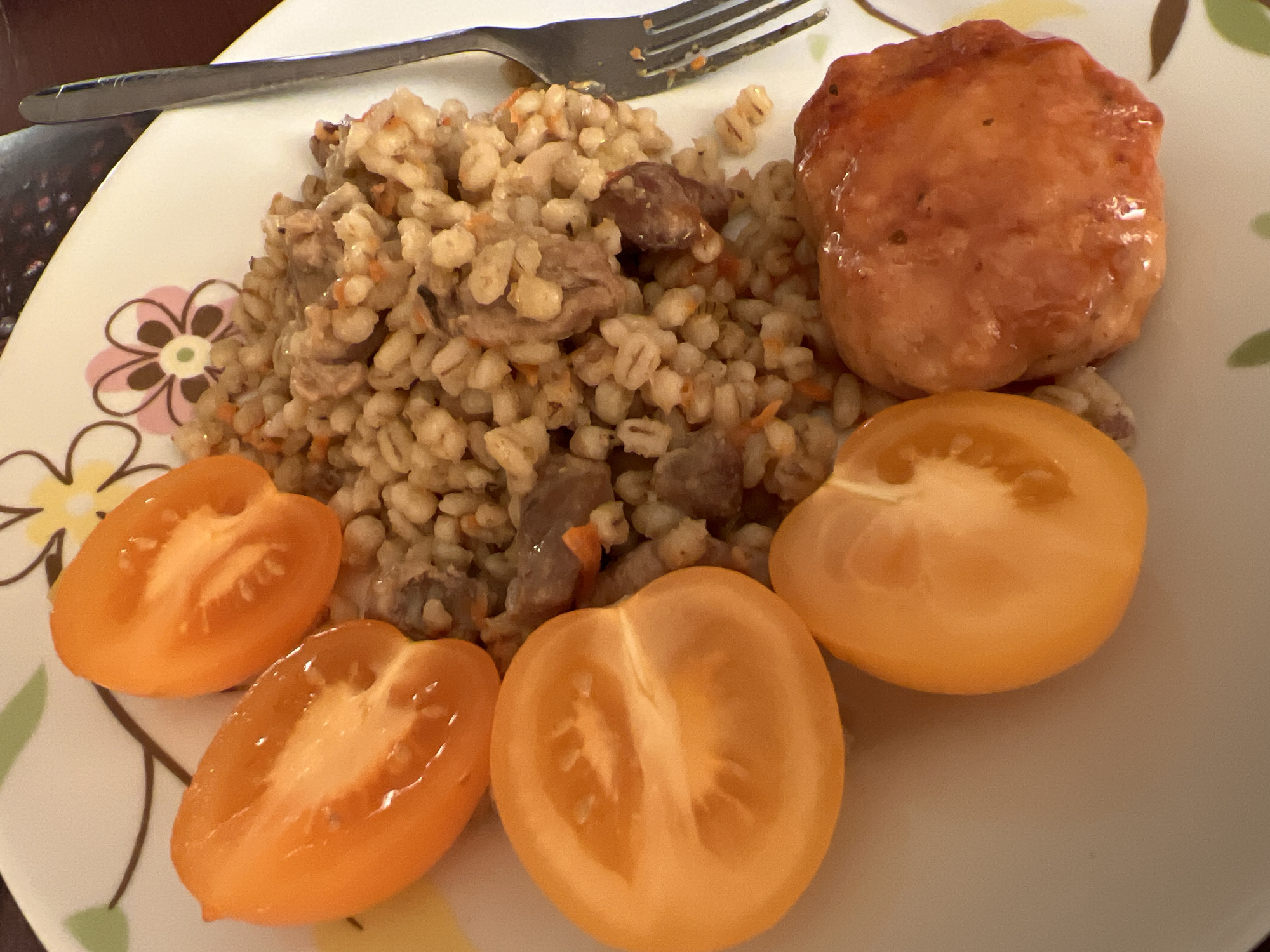 A top-down shot shows a plate of food, with four tomato halves arranged on the left side and a serving of pearl barley with meat and carrots filling the rest of the plate. A single, round meat patty sits at the bottom right of the plate. A fork rests on the right edge of the plate. The plate has a floral pattern. The food is positioned on a dark surface, suggesting it may be a table. The image appears to have been taken indoors, with artificial lighting. The location is not specified in the image itself.