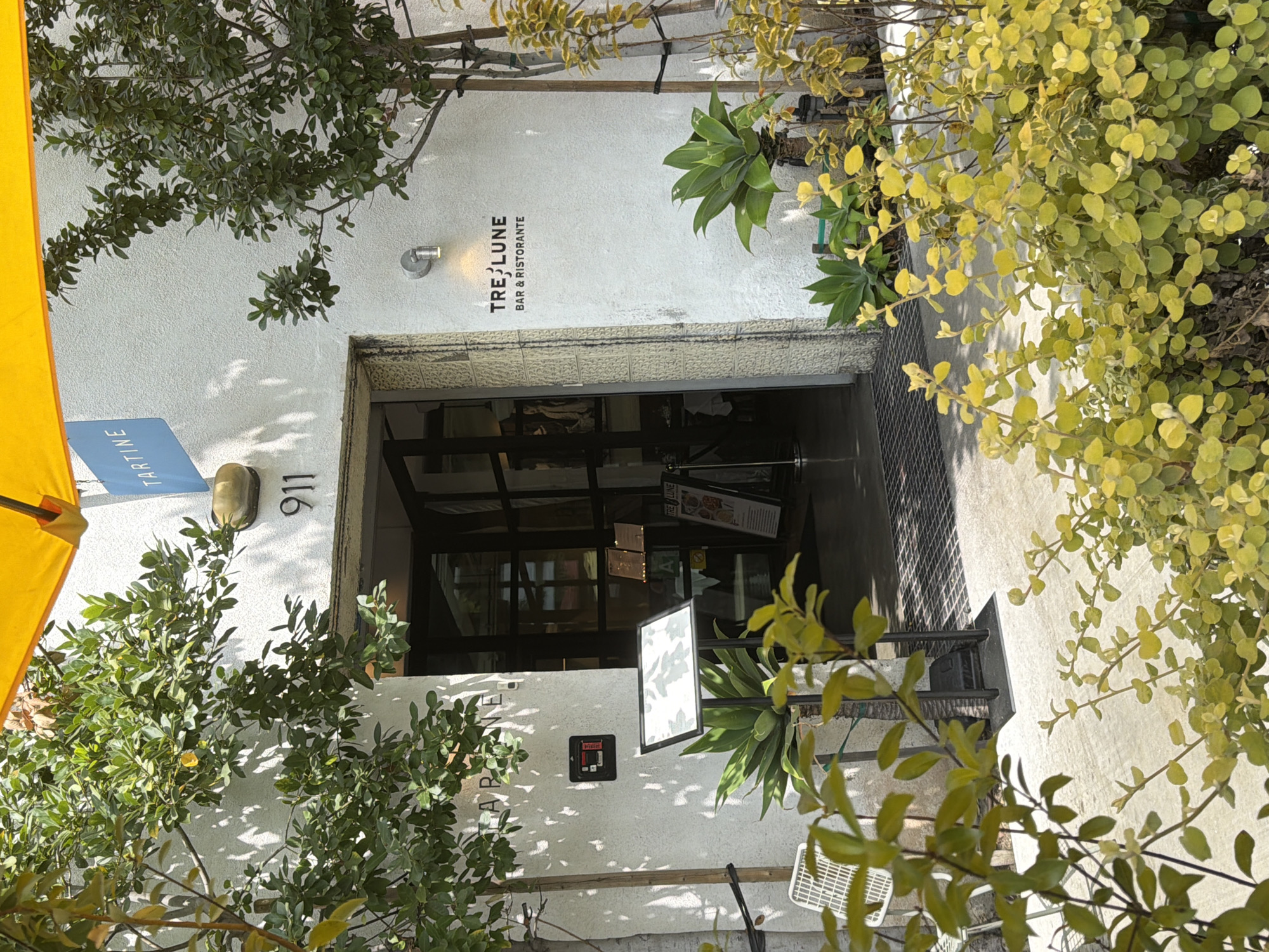 The image shows the exterior entrance to a restaurant called "TARTINE" with the address "911" visible on the wall. To the right of the entrance, another sign reads "TRE LUNE BAR & RISTORANTE". The building has a white stucco exterior and is surrounded by greenery, including trees and bushes with yellow leaves. A bright yellow awning is visible at the top of the image. The entrance itself appears to be an open doorway leading into the restaurant, with a metal grate on the ground. Several potted plants and a white outdoor chair are also present in the foreground. The scene suggests a sunny day in a vibrant urban setting, likely Los Angeles, United States of America. There are no people visible in the frame.