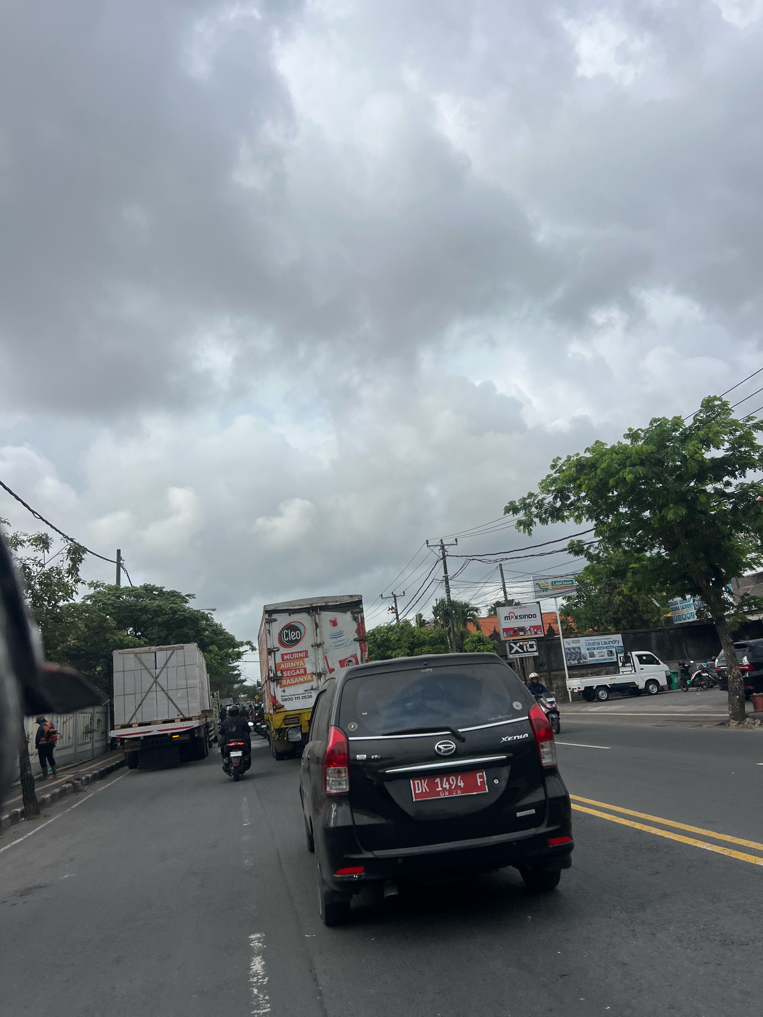 The image depicts a traffic scene in Denpasar, Indonesia, under a cloudy sky. A black car with license plate DK 1494 F is in the foreground, facing away from the viewer. Behind it, a motorcycle is visible. Further ahead, a large white truck with "Cleo" branding is in traffic, followed by another truck carrying what appears to be large, wrapped packages. On the right side of the road, there are trees, utility poles, and signs for businesses including "MAXSINDO," "X10," and "Usaha Laundry." A small white truck and several motorcycles are parked on the side. On the left side of the road, a person in an orange vest is seen walking along the sidewalk, next to a wall and trees. The road is asphalt, with yellow lane markings visible.