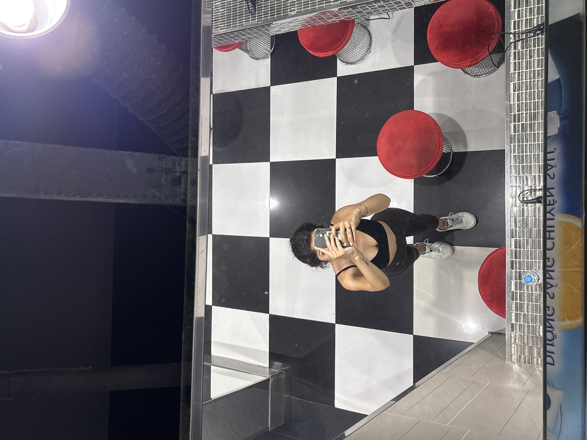 A bird's eye view shows a woman in black workout attire and sneakers taking a photo of herself in a mirror. The floor is covered in a checkerboard pattern of black and white tiles. Red stools are positioned around the room. There is a light fixture and ventilation in the ceiling. There is also a beverage cooler with an orange displayed behind the woman. The location is likely in Đà Nẵng, Vietnam.