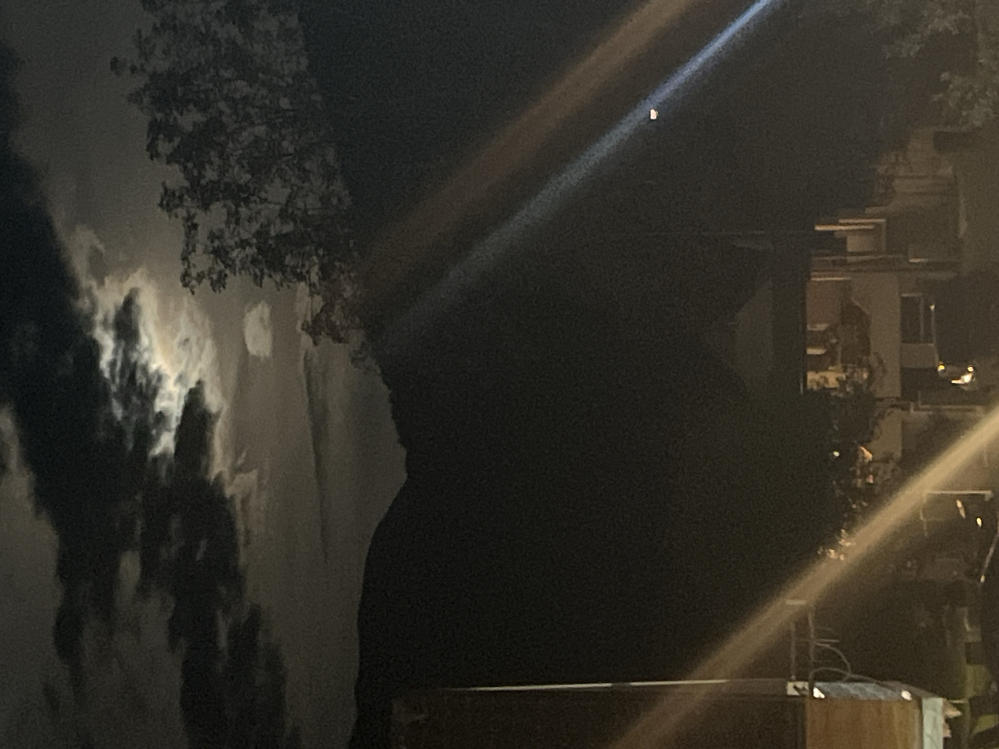 The image shows a night scene. On the left are clouds, partly illuminated. A structure and trees are present in the middle-ground. A light beam extends diagonally across the frame, originating near the top. Another angled light beam appears on the right side of the image. Buildings and trees can be seen in the background on the right. The image is dark, with varying degrees of illumination from the light sources. There is no visible evidence that shows this is located in the city of Polja, Montenegro.