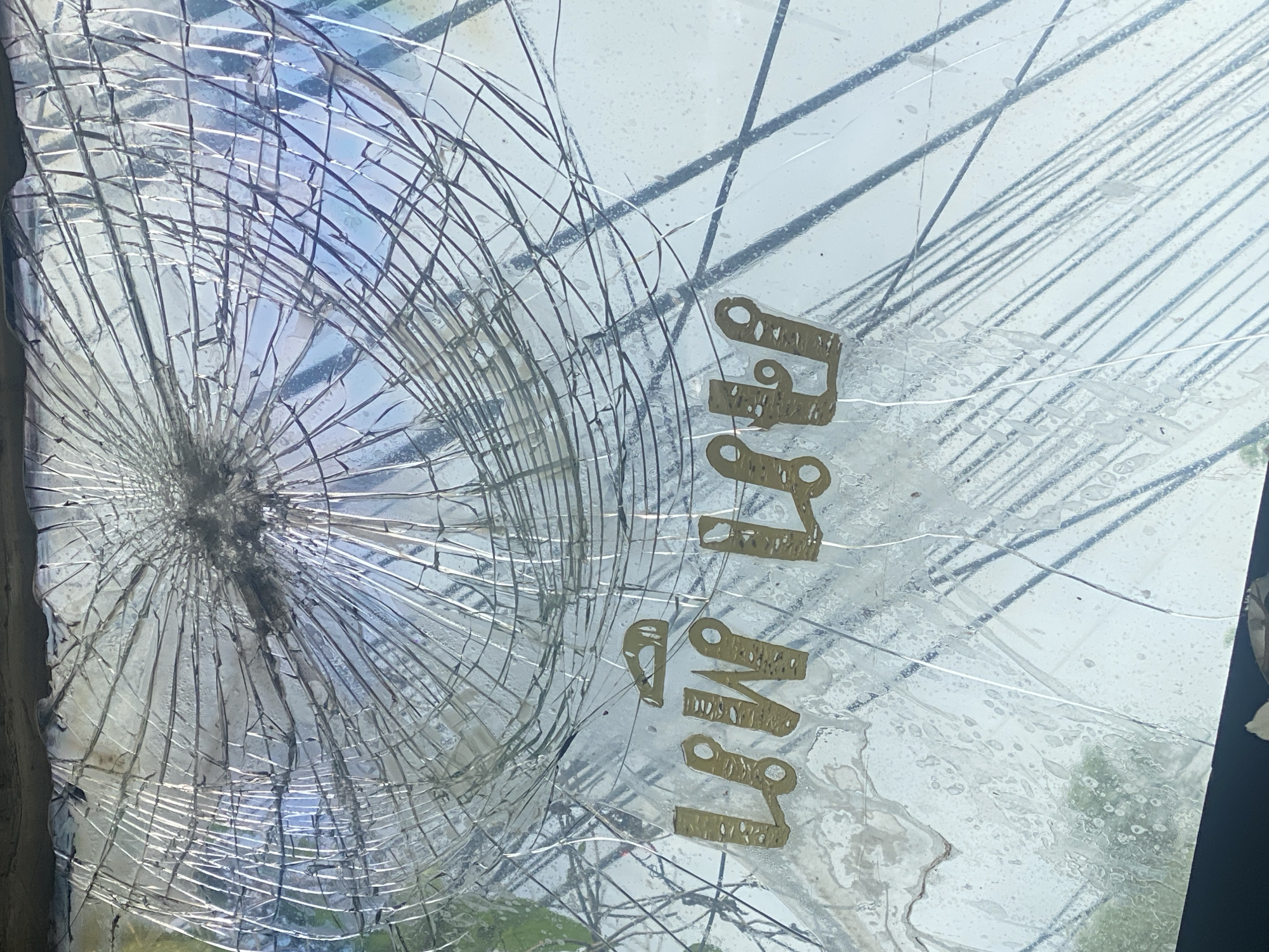 A severely cracked glass surface obscures the view. The center of the cracking is on the left side. Partially visible through the glass and the cracks are power lines and trees, suggesting an outdoor setting. The Thai word "ท่งศาลา" (Thong Sala) is printed vertically in gold lettering on the glass. The state of the glass and the presence of the word indicate that this is a broken window or advertisement in Thong Sala, Thailand.