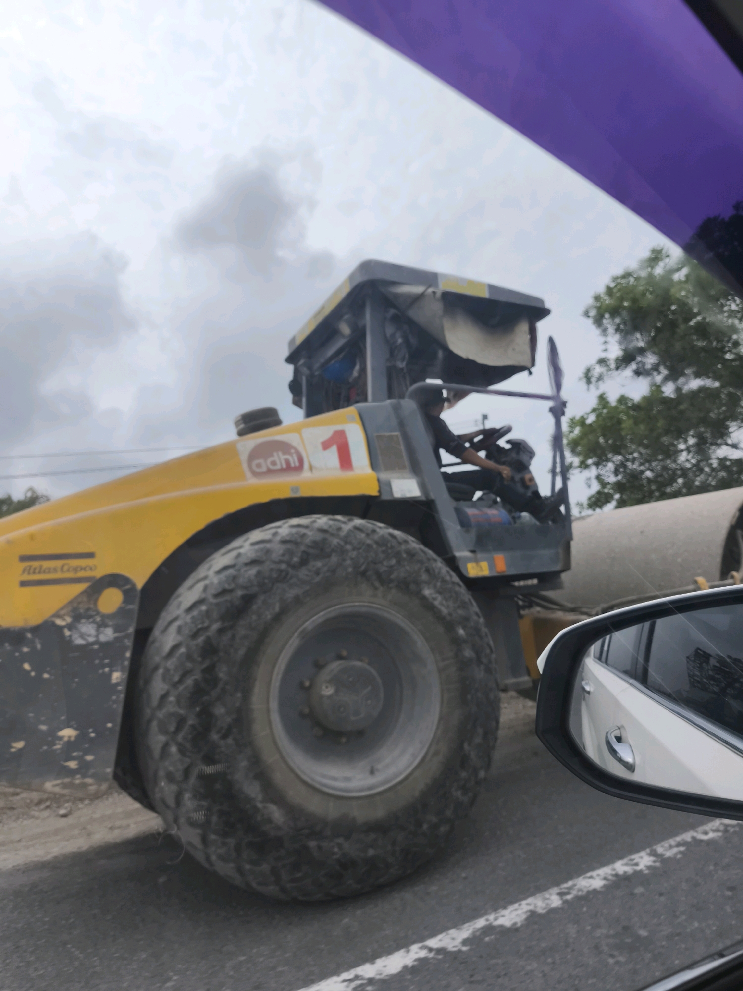 A yellow Atlas Copco vibratory roller, numbered "1" and with the logo "adhi", is shown on a road construction site. The roller has a large, treaded tire. A person wearing black clothing is operating the machine from an open cab. In the background, a large concrete cylinder is visible, along with trees and a cloudy sky. A side mirror of a white car is in the foreground, reflecting the construction site and a building. The image appears to be taken from inside a vehicle, with a purple tinted window visible at the top. The setting is identified as Mayapati, Indonesia.