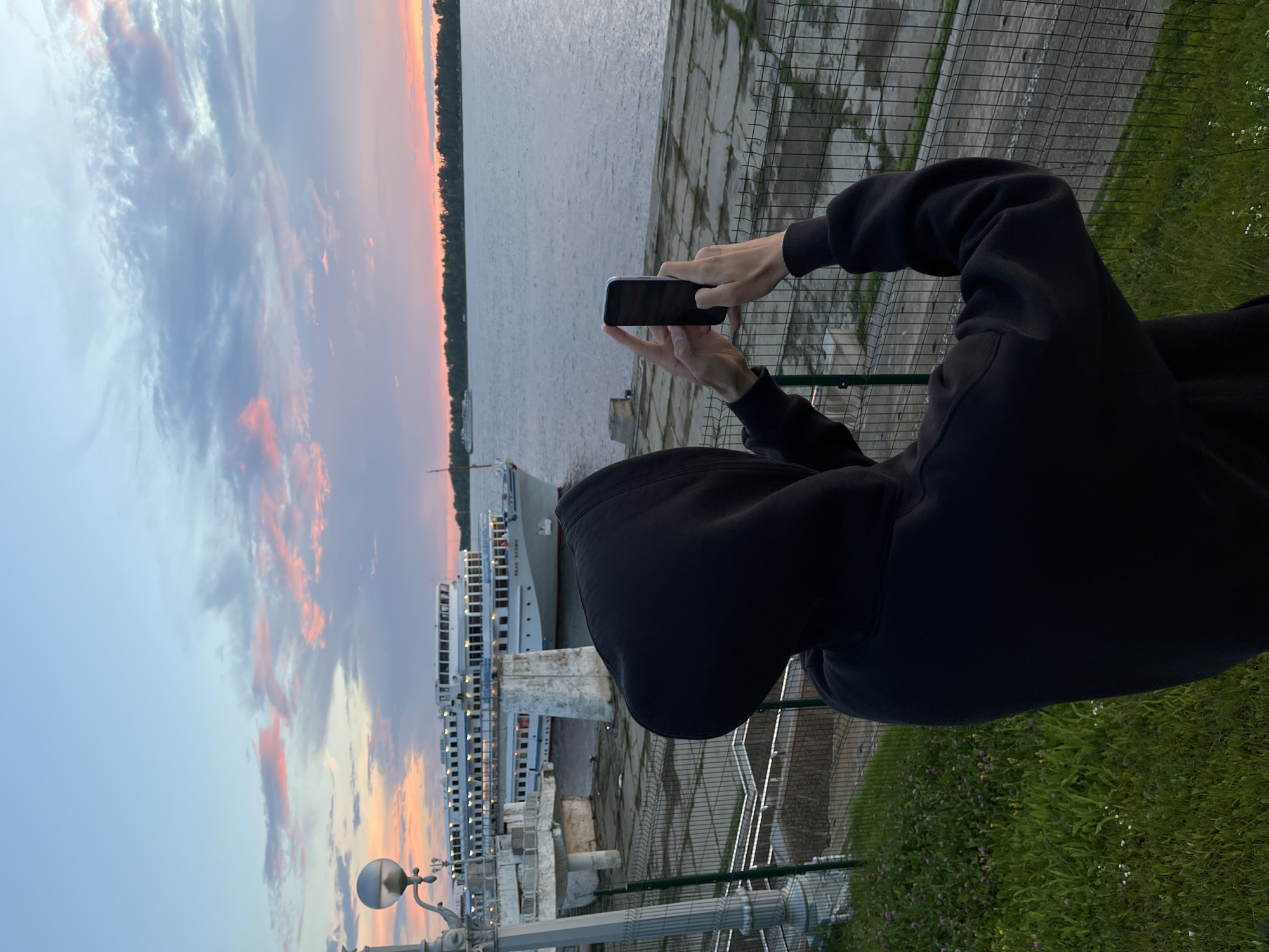 This media file captures a person from behind, wearing a black hooded sweatshirt, holding a smartphone and taking a photo. The scene is a waterfront setting, likely a pier or embankment, with a body of water extending into the background. A large white ship is docked at the pier. The sky displays a colorful sunset with hues of orange, pink, and blue in the clouds, suggesting it is dusk. The ground area near the person is covered in grass with small wildflowers, and a metal fence is visible. The overall atmosphere appears serene and picturesque, with the subject engaged in documenting the scenic view. The location is identified as Novokuybyshevsk, Russia. No visible text is present in the image.