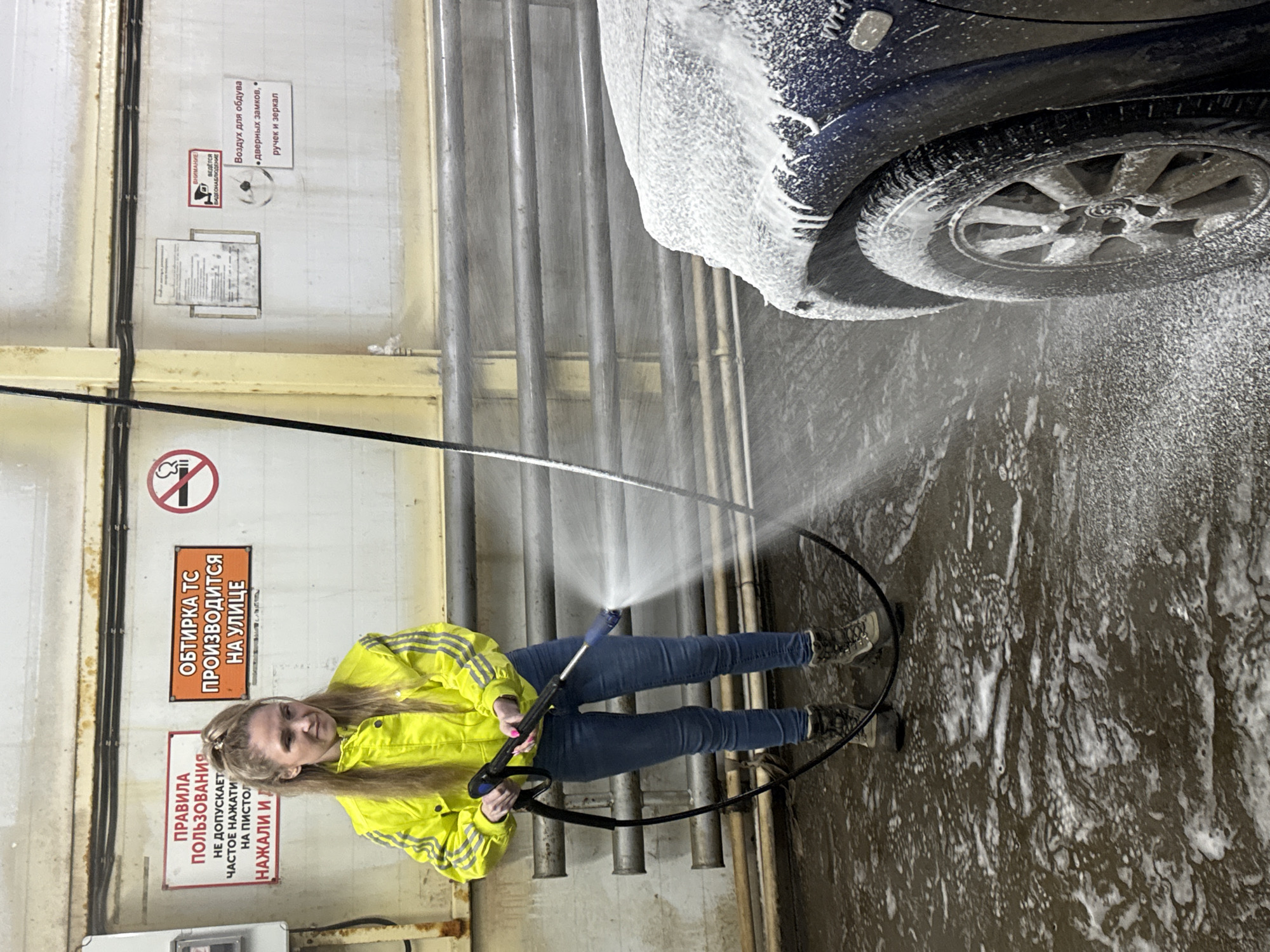 The image depicts an indoor self-service car wash bay in Chaikovsky, Russia. A woman, appearing to be in her late 20s to early 30s, stands centrally, operating a high-pressure washer. She has long, light brown hair tied in a high ponytail and is wearing a bright yellow jacket with gray stripes on the sleeves, blue denim jeans, and dark gray hiking boots. She holds the black and blue pressure washer gun with both hands, directing a stream of foamy water towards the front right quarter of a dark blue vehicle, which is partially visible on the right side of the frame. The vehicle's front fender, a portion of the hood, and its wheel are extensively covered in white soap suds.
The floor of the bay is wet and covered in white soap foam. In the background, light-colored walls feature multiple signs in Russian: one red sign on the left provides "RULES OF USE," prohibiting "frequent pressing on the pistol." An orange sign in the center states that "VEHICLE WIPING/DRYING IS DONE OUTSIDE." To the right, a circular "no smoking" symbol is visible. Further right, a sign indicates "ATTENTION! VIDEO SURVEILLANCE IS CONDUCTED," and another specifies "Air for blowing • door locks, • handles and mirrors." Horizontal metal pipes or rails are mounted along the walls. A black hose connected to the pressure washer lies coiled on the wet floor around the woman's feet.