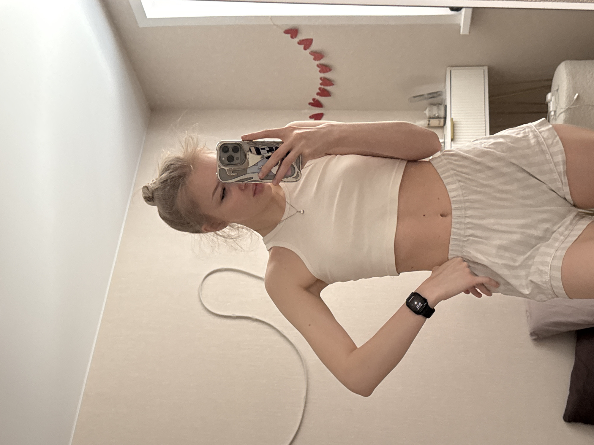 A young woman is taking a selfie in what appears to be a bedroom or apartment. She is wearing a white crop top and striped shorts, and has a black smartwatch on her left wrist. Her hair is styled in a bun. She is holding a smartphone with a decorative case in her left hand, obscuring part of her face. In the background is a mirror with small red heart decorations above it, a white cabinet, and part of a bed with pillows. The location is implied to be Perm, Russia, though there is no visual confirmation.
