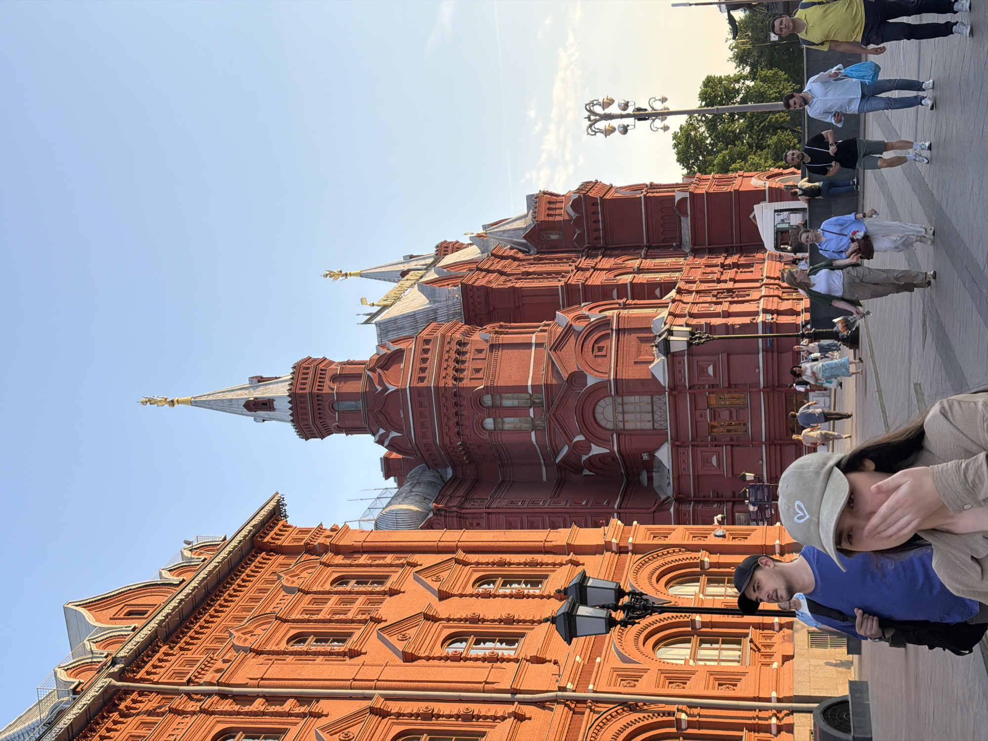 The image depicts the State Historical Museum on Red Square in Moscow, Russia. The museum's distinctive red brick facade is prominent, featuring arched windows and decorative architectural elements. Several individuals are present in the foreground and along the sidewalk adjacent to the museum, including a couple in the bottom right, with the woman covering her mouth. Others are walking along the square, in varied attire. A vintage street lamp stands out near the bottom, and other similar lamps are visible further down the street. The sky is clear and blue.