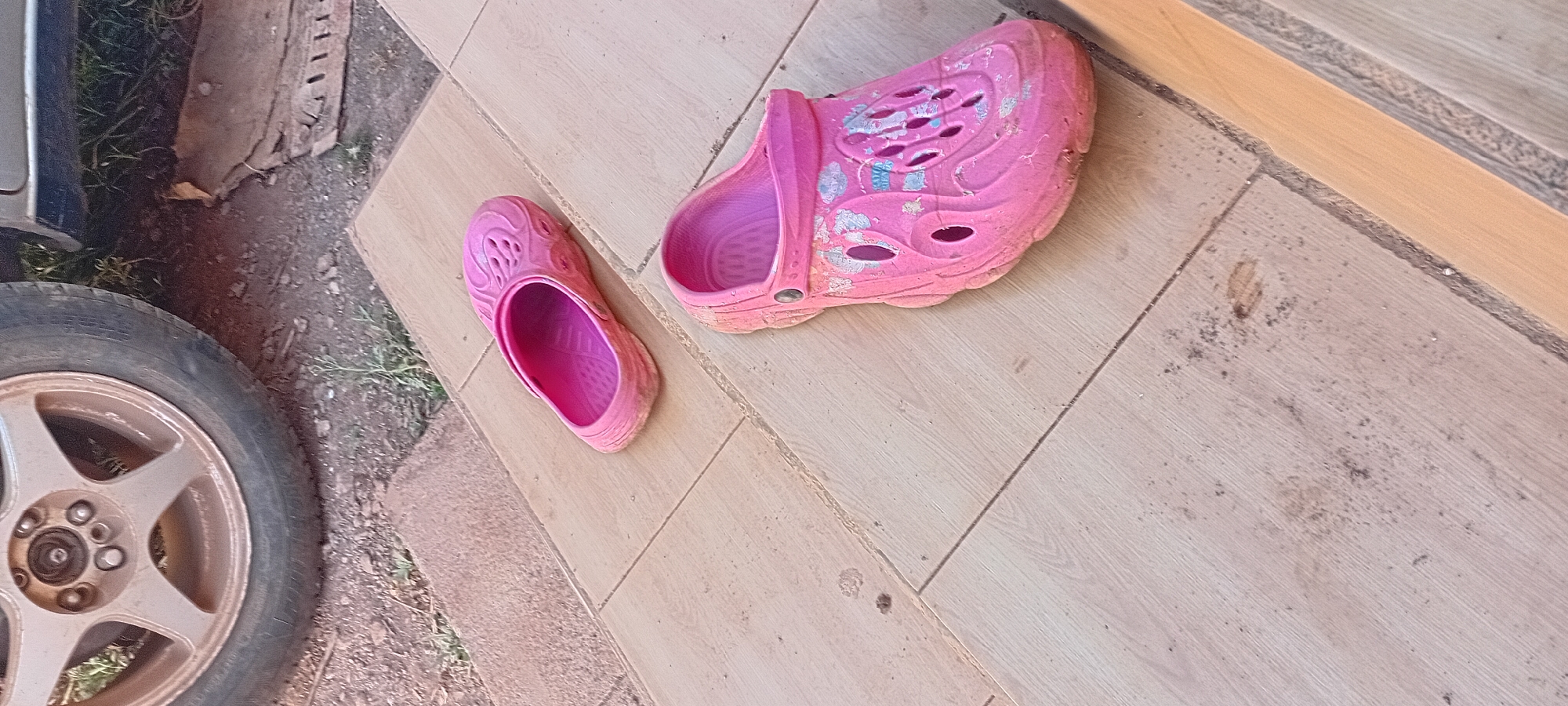 The image depicts two pink children's clogs on a tiled outdoor floor. The clogs are worn and show signs of dirt and scuffing. One clog is positioned to the left, and the other is to the right and slightly further down. In the background, a portion of a car wheel and tire is visible, along with dry grass and a rough, mat-like object on the ground. The overall environment suggests an outdoor setting, possibly a residential area in Kijabe ward, Kenya, given the context. The lighting indicates daytime. No individuals are present in the frame.