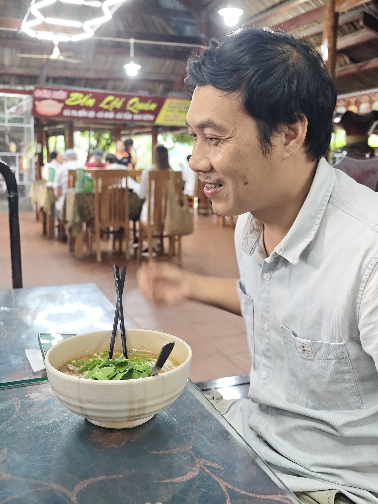 A man is sitting at a table in an outdoor restaurant in Nam Khanh Vinh, Vietnam. He is wearing a light blue collared shirt and is looking to his right, smiling. In front of him is a bowl of pho with chopsticks and a spoon. The restaurant appears to be busy, with other patrons visible in the background. The ceiling is made of wood and corrugated metal, and there are hanging lights. The table is made of glass and metal.