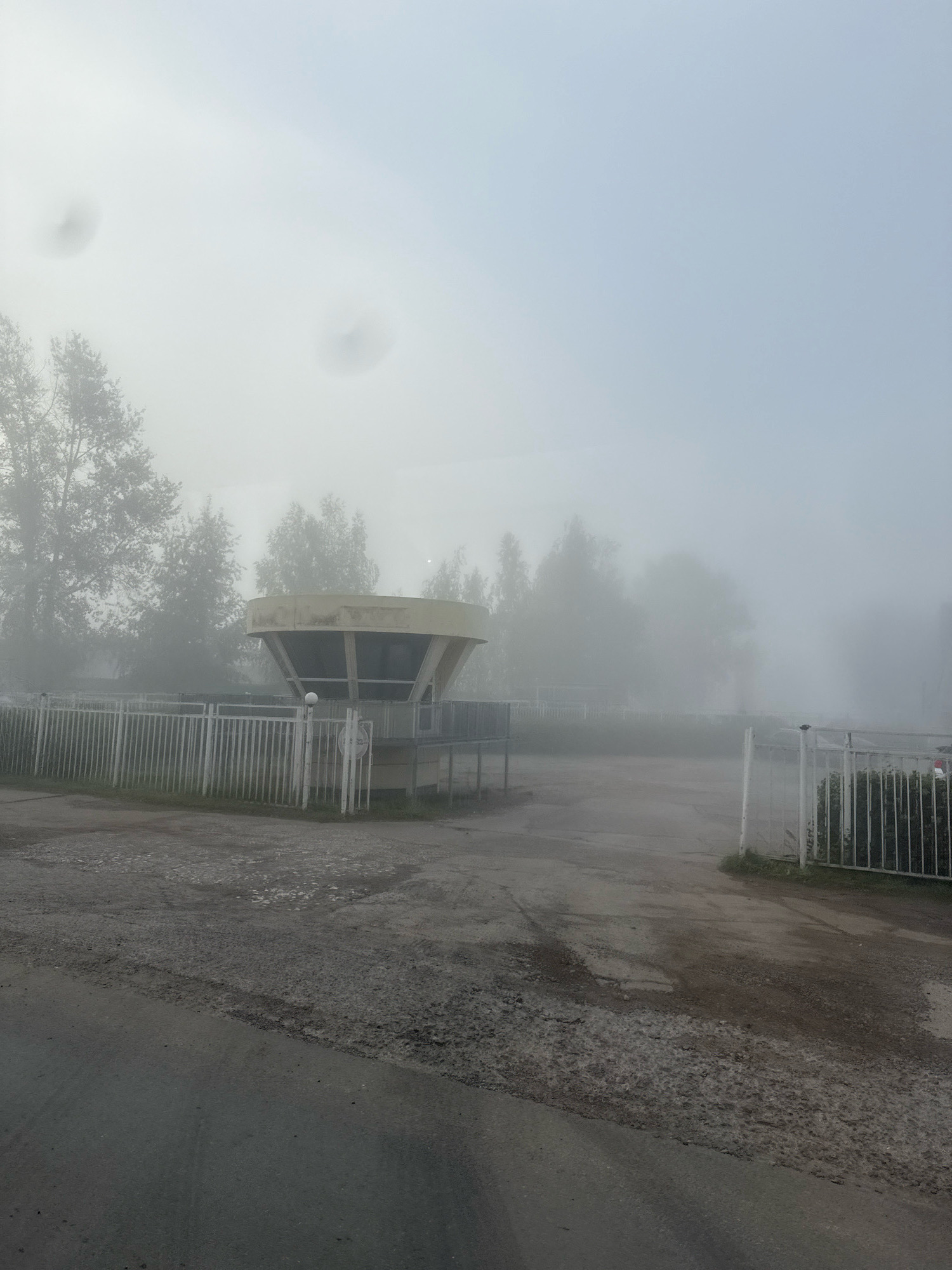 In Chaikovsky, Russia, a photograph captures a foggy morning scene near a building with a circular, tiered roof structure supported by pillars. The building appears to be a bus stop or a small pavilion. A white metal fence with vertical slats runs along the front of the property, and an opening in the fence suggests an entrance. Beyond the fence, trees are visible, shrouded in the dense fog, making their outlines indistinct. The ground in front of the fence is paved with asphalt that shows signs of wear and damage, with patches of gravel and uneven surfaces. The sky is overcast and pale blue, contributing to the overall muted and atmospheric quality of the image. Two indistinct dark circular shapes are visible in the sky, possibly lens flares or atmospheric phenomena.