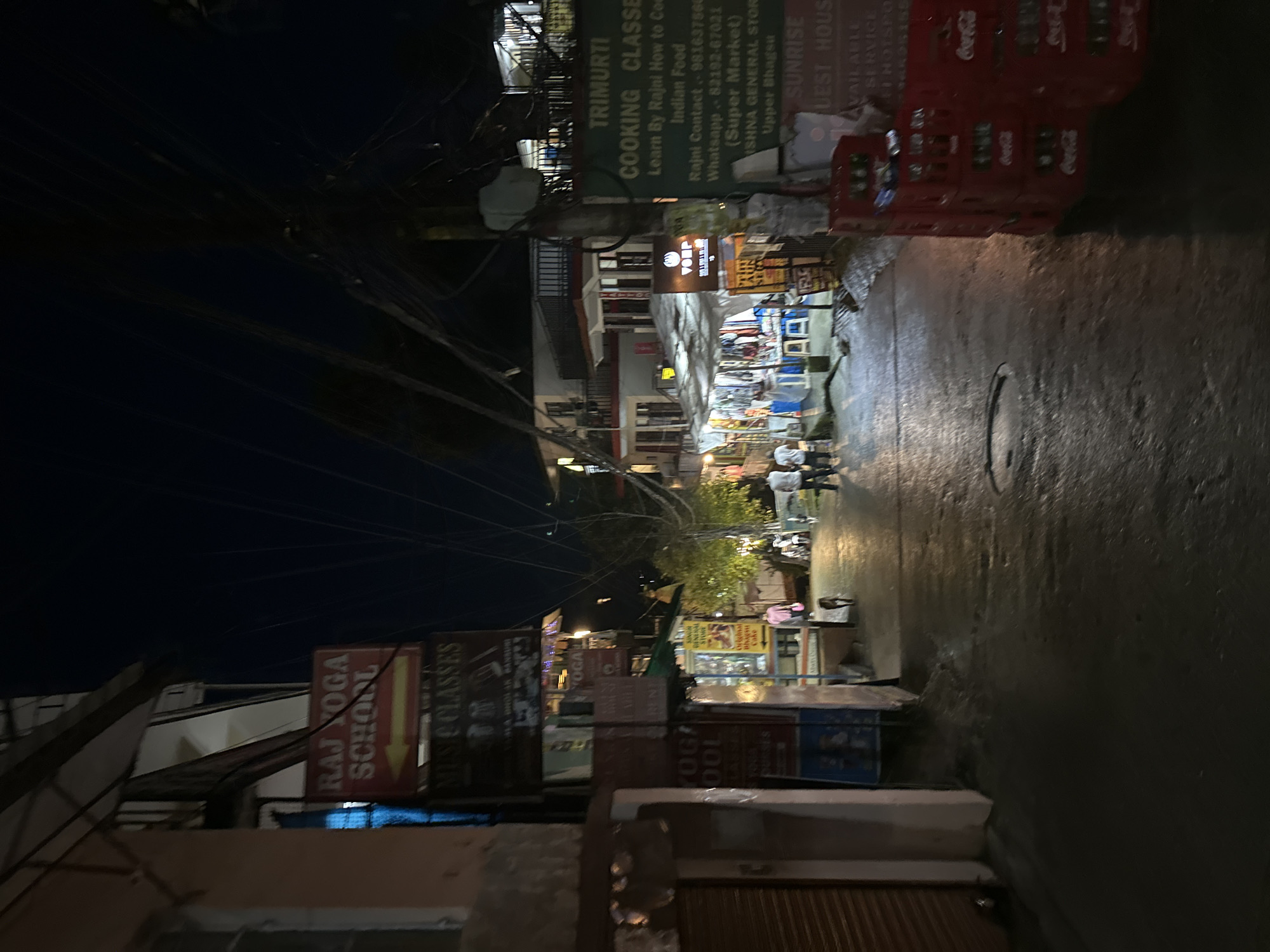 The image depicts a narrow street at night, illuminated by artificial lights. On the left side of the street, a building with a peach-colored facade features signage for "RAJ YOGA SCHOOL" and "MUSIC CLASSES." Further down the street, a row of market stalls with white canopies are set up, displaying various items. A few people are visible walking along the street, including two individuals in white shirts and pants, and one person in a pink shirt. A dog is also present. The street surface appears wet and reflective. On the right side of the street, a green sign for "TRIMURTI COOKING CLASSES" is prominent, along with a sign for "SUNRISE GUEST HOUSE." Stacks of red Coca-Cola crates are piled near the right edge of the image. The sky above is dark, with a dense network of electrical wires visible.