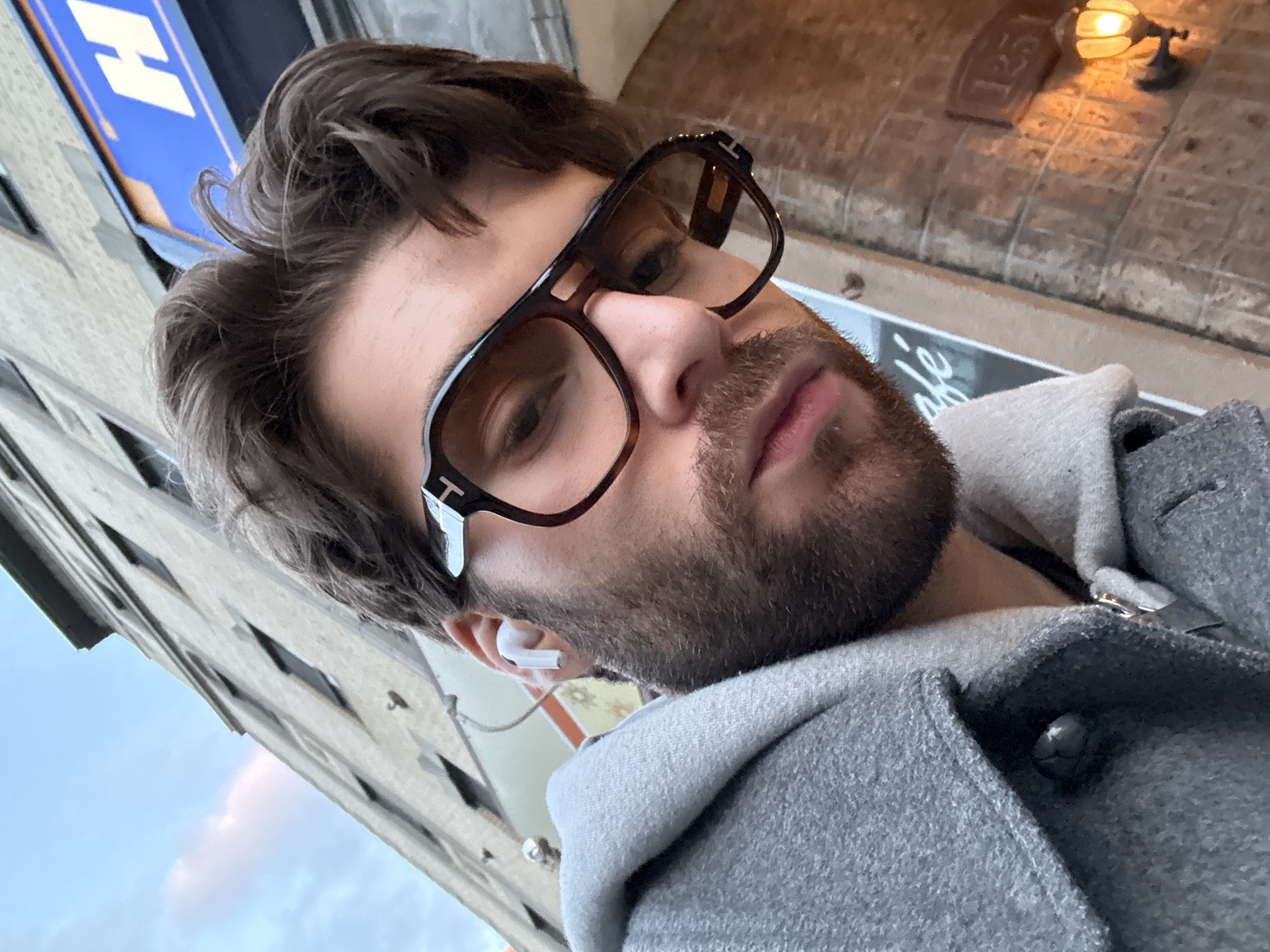 A close-up, low-angle self-portrait of a man with dark hair and a beard, wearing dark-rimmed glasses and a gray hooded jacket. He has a wireless earbud in his left ear. Buildings are visible in the background, including one with a blue sign. A brick building with a lit lamp is also visible. The location is likely in Montreal, Canada.