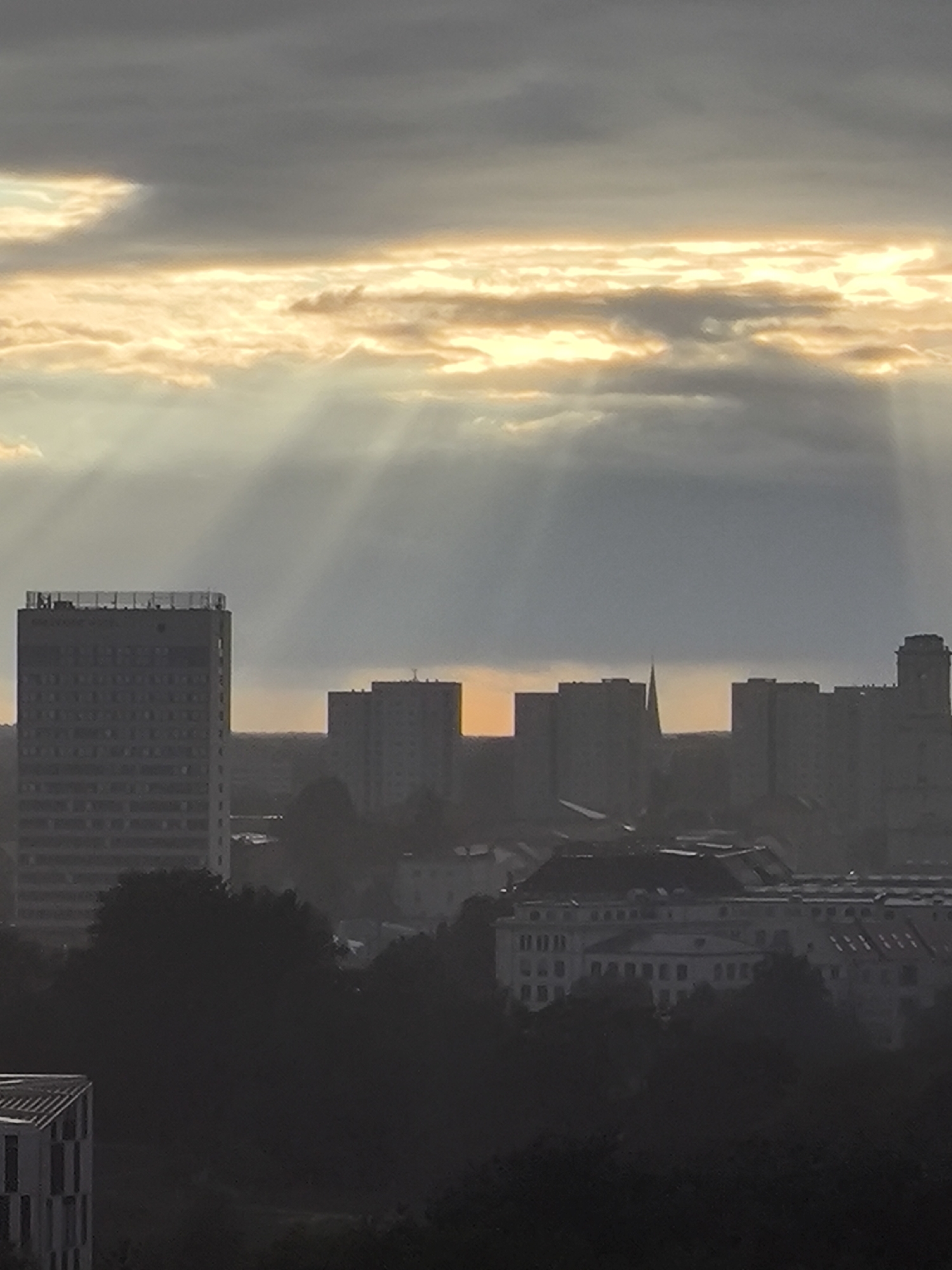 The image depicts a cityscape during what appears to be either sunrise or sunset, with dramatic sunbeams breaking through clouds. In the foreground, silhouetted trees and buildings obscure much of the lower portion of the scene. A notable structure on the left is a tall, rectangular building with a visible rooftop structure. Further back, a series of apartment buildings, characteristic of the Plattenbau style common in East Germany, are silhouetted against the hazy sky. A pointed spire, potentially belonging to a church or a tower, is visible amongst these buildings. The overall atmosphere is somber and somewhat diffused due to the atmospheric conditions. This scene is consistent with views found in Potsdam, Germany, possibly overlooking residential areas with high-rise apartment blocks.