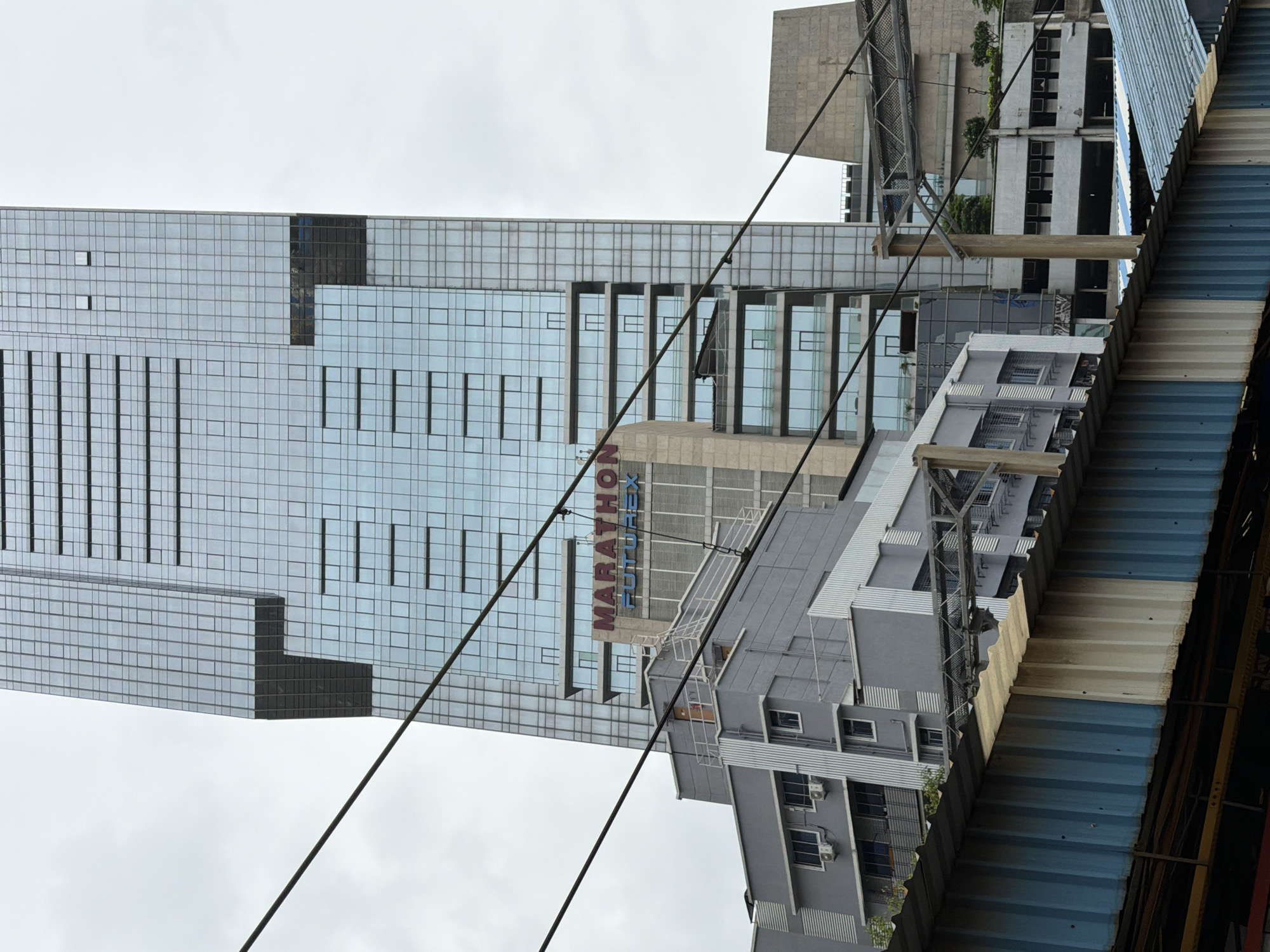 The image shows a daytime, low-angle shot of the Marathon Futurex building in the Lower Parel area of Mumbai, India. The building is a modern skyscraper with a glass facade, reflecting the cloudy sky. In the foreground, there is a corrugated metal structure, likely part of a shelter or barrier. Overhead power lines crisscross the frame, partially obscuring the view of the buildings. To the left of the main skyscraper, a multi-story gray building is visible, featuring balconies and air conditioning units. The Marathon Futurex sign is prominently displayed on a beige section of the skyscraper. Other buildings and infrastructure, including what appears to be a bridge or elevated walkway, are visible in the background. The scene is urban and industrial, with a focus on modern architecture and infrastructure. There are no people visible in the image.