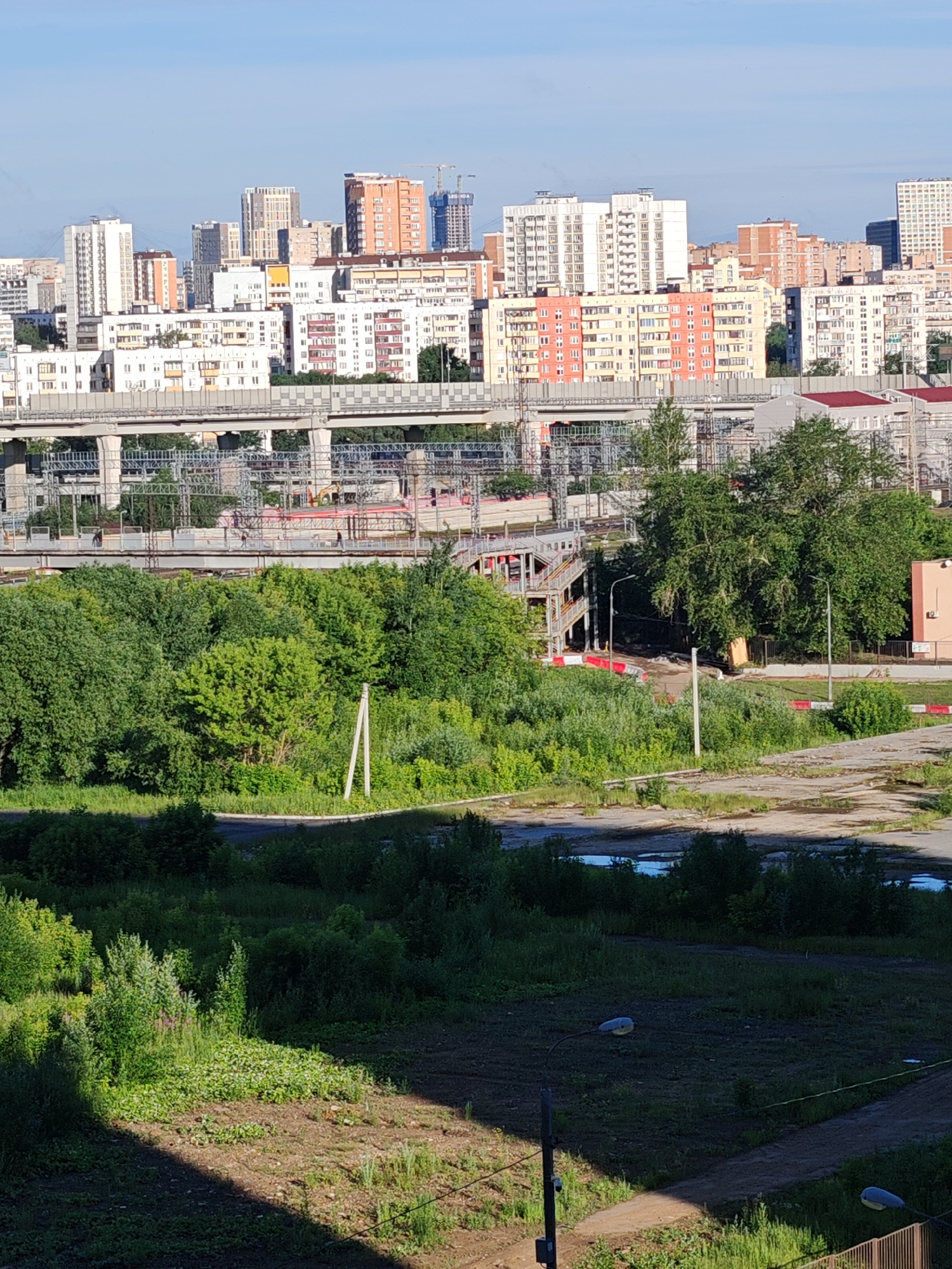 The image depicts a cityscape with a clear blue sky. In the foreground, there is a patch of bare earth with some sparse vegetation and a shadow cast from an unseen object. Beyond this, a road or path is visible, lined with greenery on either side. Further back, a dense expanse of lush green trees and bushes occupies the mid-ground. Above the trees, a large elevated railway or road structure stretches across the frame, supported by concrete pillars and featuring extensive scaffolding and construction elements. Beyond this structure, a collection of multi-story buildings, ranging in color from white and cream to orange and red, forms the urban skyline. Some of these buildings appear to be residential apartments, while others might be office buildings. Construction cranes are visible among the taller structures, indicating ongoing development. The scene is illuminated by natural daylight, suggesting it is daytime.