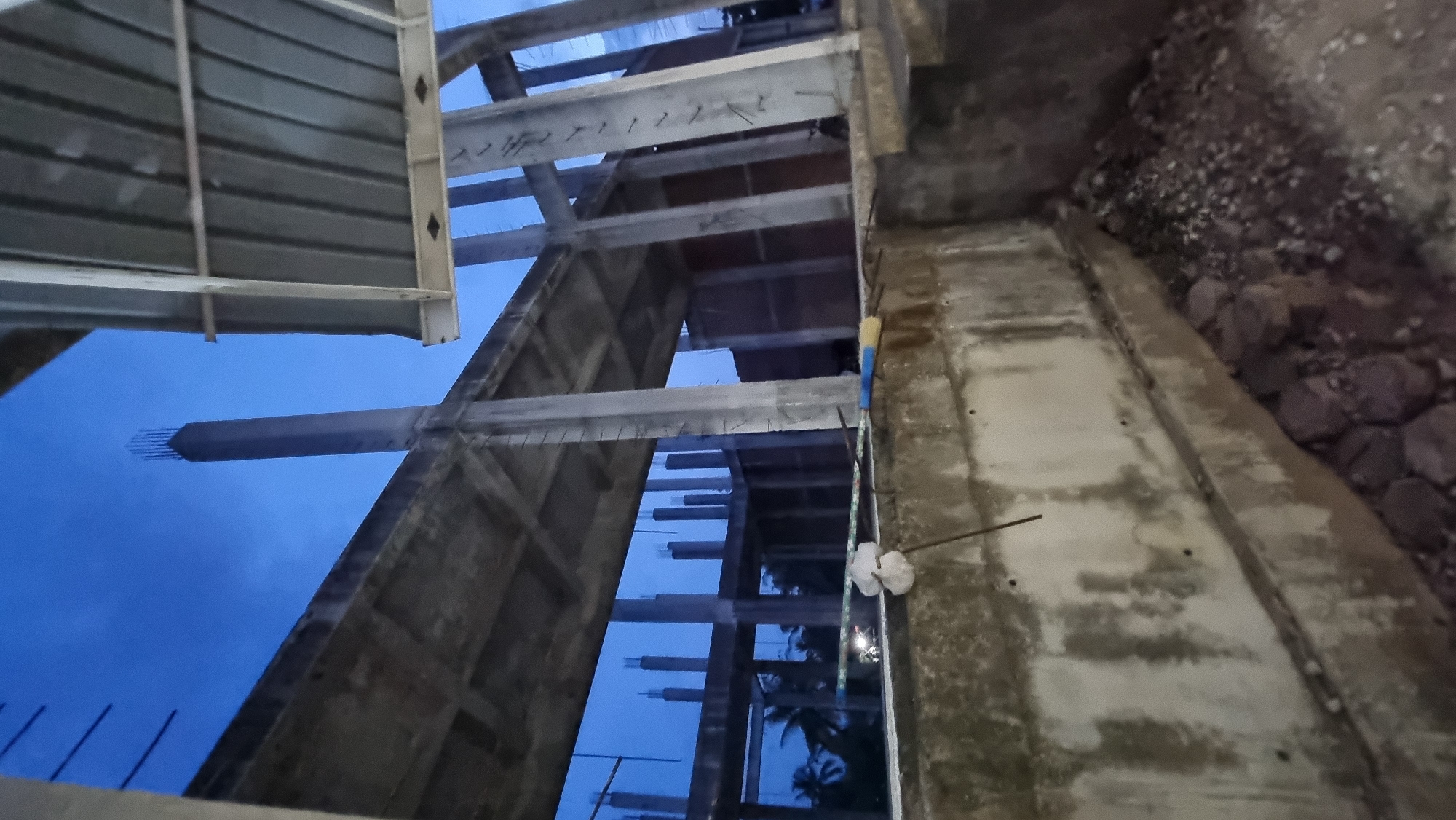 A construction site in Manetunong, Indonesia, shows the skeletal framework of a building under a twilight sky. Concrete pillars and beams form the primary structure, with rebar protruding from some of the upright columns. In the foreground, a concrete wall with visible stains and marks runs horizontally across the image. Scattered on the ground near this wall are small rocks and debris. In the background, several unfinished concrete pillars are visible, suggesting the ongoing construction of multiple levels. Palm trees can be seen in the distance through the open structure, indicating a tropical or subtropical environment. The overall scene depicts the early stages of building construction at dusk.